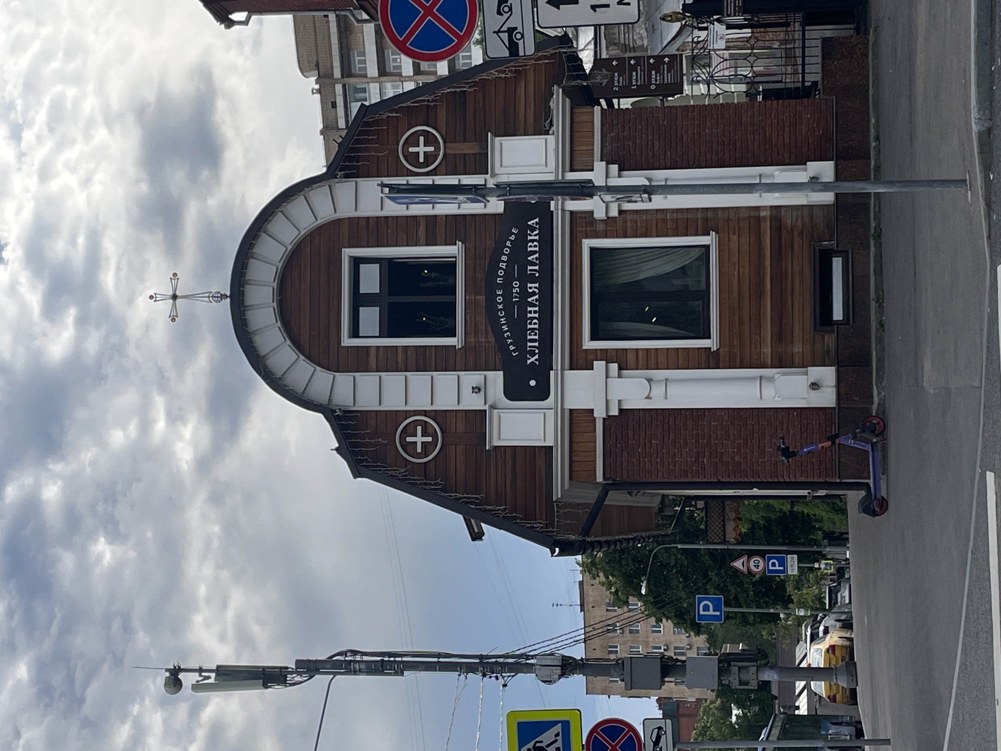 The image depicts a street scene in Moscow, Russia, featuring a distinctive wooden building with a dark sign that reads "ГРУЗИНСКОЕ ПОДВОРЬЕ 1750 ХЛЕБНАЯ ЛАВКА". This translates to "Georgian Courtyard 1750 Bakery Shop". The building has a mansard roof topped with a cross and white decorative molding around the arched windows. A cell tower stands prominently to the left of the building, equipped with antennas and various fixtures. Several traffic signs are visible, including a pedestrian crossing sign, a "no parking" sign, and speed limit signs. A purple electric scooter is parked on the sidewalk in front of the building. In the background, apartment buildings and trees can be seen. The sky is overcast with clouds. Cars are parked and driving on the street.