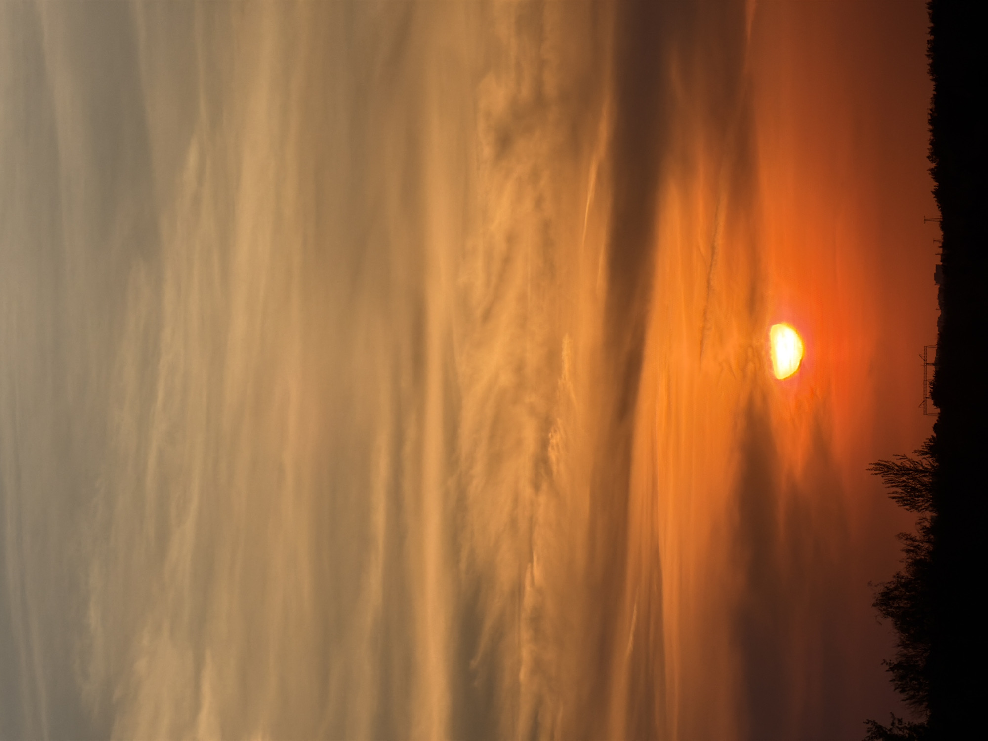 The image depicts a sunset in an unspecified location in the city of Unknown, Unknown. The sun is partially visible near the horizon, obscured by clouds. The sky transitions from orange near the sun to grey further away. A dark treeline and a few power lines are visible on the right. There are no people visible in the image.