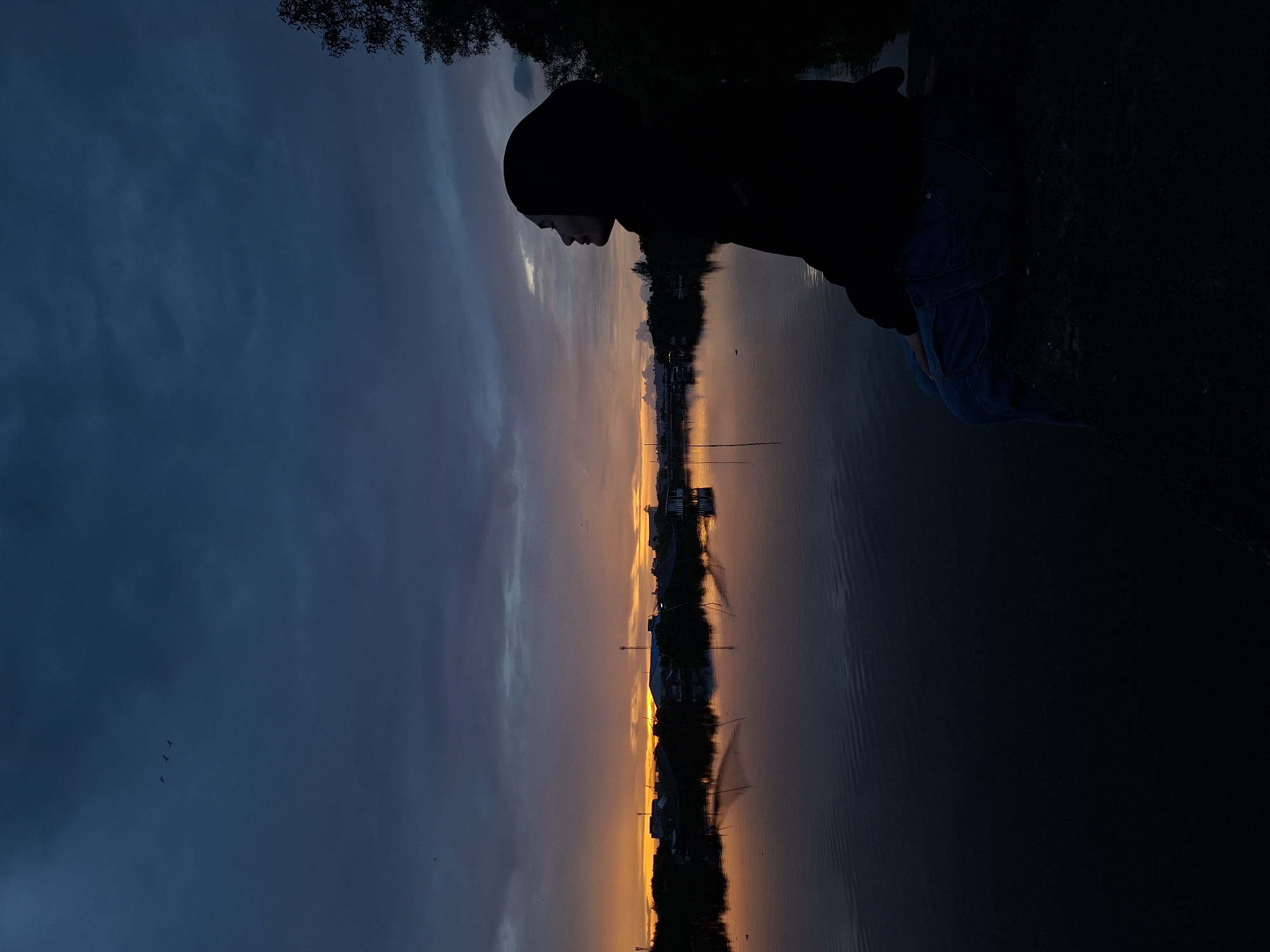 A nighttime outdoor scene shows a person, silhouetted and facing left, standing in front of a body of water. A small town is visible across the water with lights and buildings reflecting on the surface. The sky is a mix of blue and orange from a sunset or sunrise. In the dark blue portion of the sky, a few birds are flying. Some trees and vegetation are visible on the bank near the person. The location is presumed to be Lhokseumawe, Indonesia.