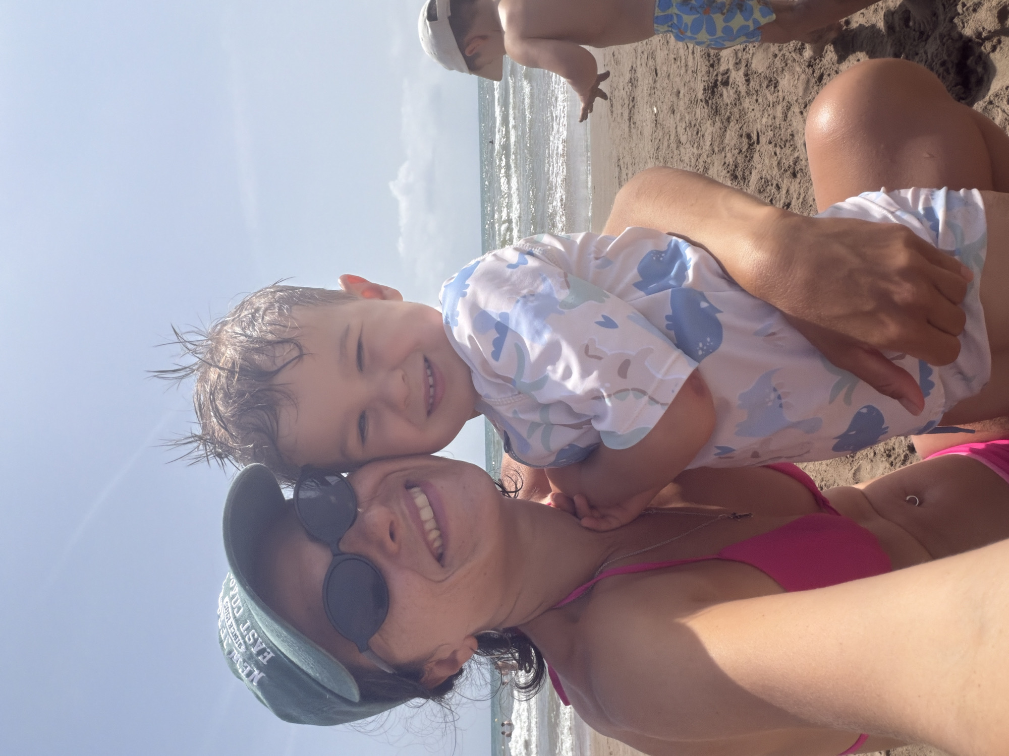This media file shows a close-up, low-angle selfie of two people on a sandy beach, with the ocean and sky in the background. The foreground features a smiling woman wearing sunglasses and a green baseball cap, and a young child with a playful expression, their heads close together. The child is wearing a light-colored shirt with a blue dinosaur pattern. The woman is wearing a pink bikini top. Another person, a child, is visible in the upper background, wearing a light-colored cap and patterned shorts, with their back to the camera.
The scene appears to be a sunny day, judging by the bright light and shadows, likely in the late morning or afternoon. The water suggests a beach location, and the overall atmosphere is relaxed and happy, indicated by the smiles of the subjects. The sand is visible in the foreground and mid-ground. The sky is bright blue and clear, suggesting good weather. No specific location cues are discernible from the image itself.
Visible text on the woman's baseball cap reads "NEW EAST COLO SINCE 1995".