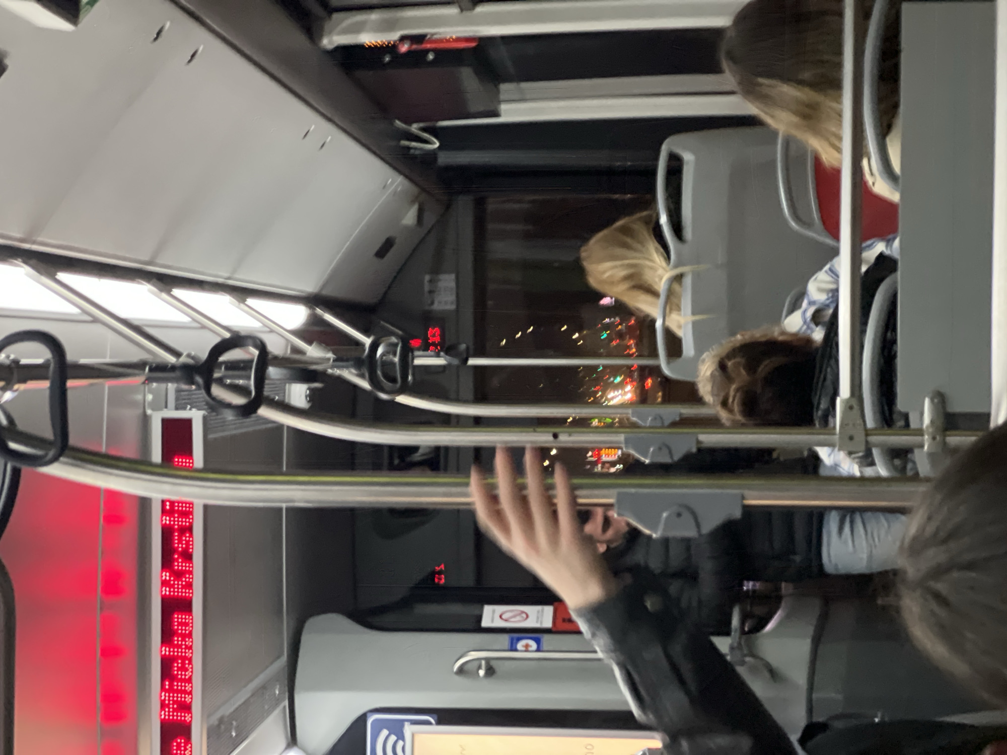 The image shows the interior of a public transportation vehicle, likely a bus, in Palilula Urban Municipality, Serbia. Several passengers are visible, seated and standing, with a hand reaching toward an overhead bar. The outside is visible through the windows, showing lights at night. Electronic displays with red lights are visible inside the vehicle.