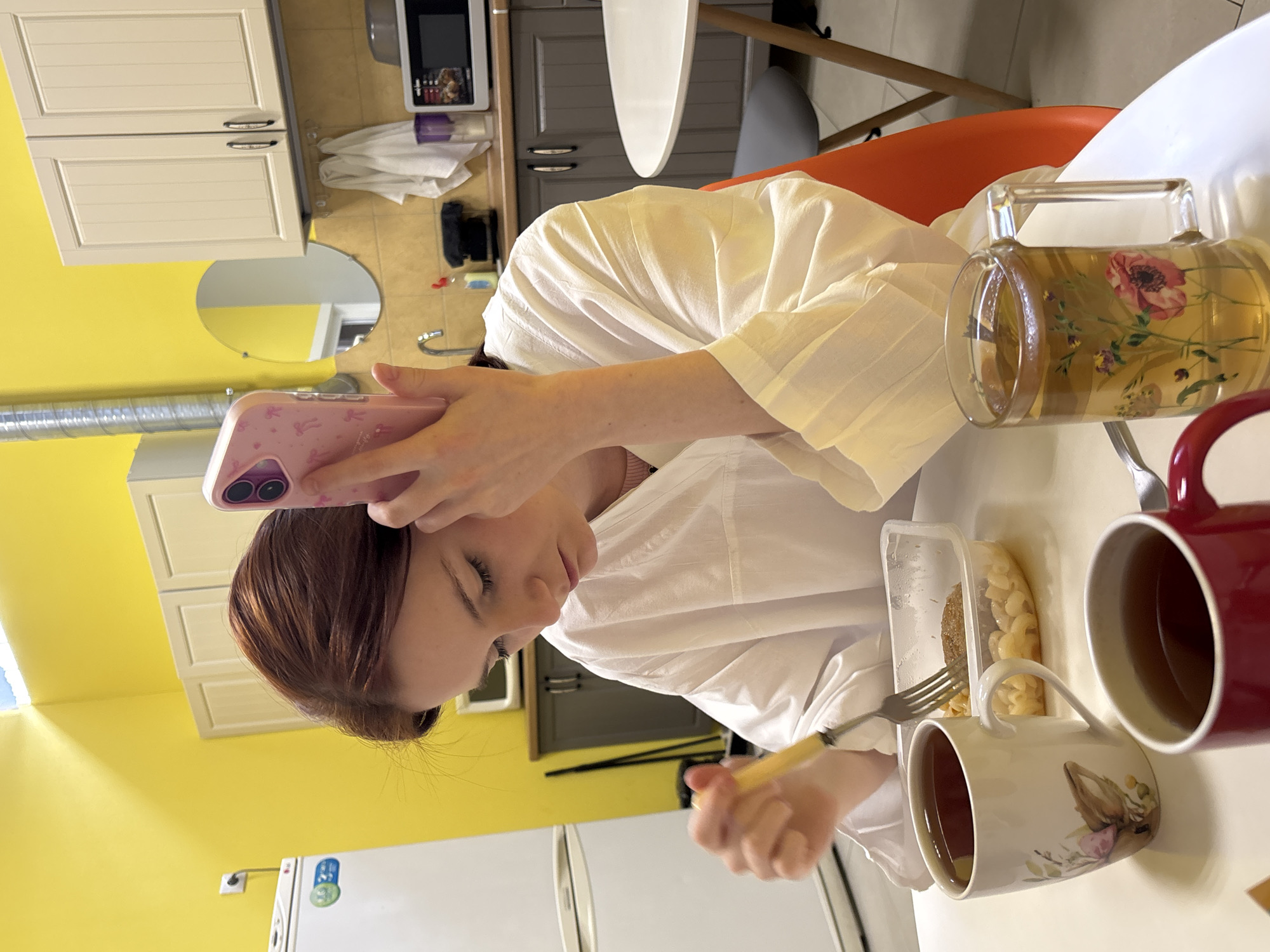 A woman in a white shirt is holding a pink cell phone while eating at a table in a kitchen. She has reddish-brown hair. On the table, there is a container of food, a mug, and a glass teapot filled with tea. The kitchen has yellow walls and white cabinets. The image does not contain sufficient information to determine the location in Saint Petersburg, Russia.
