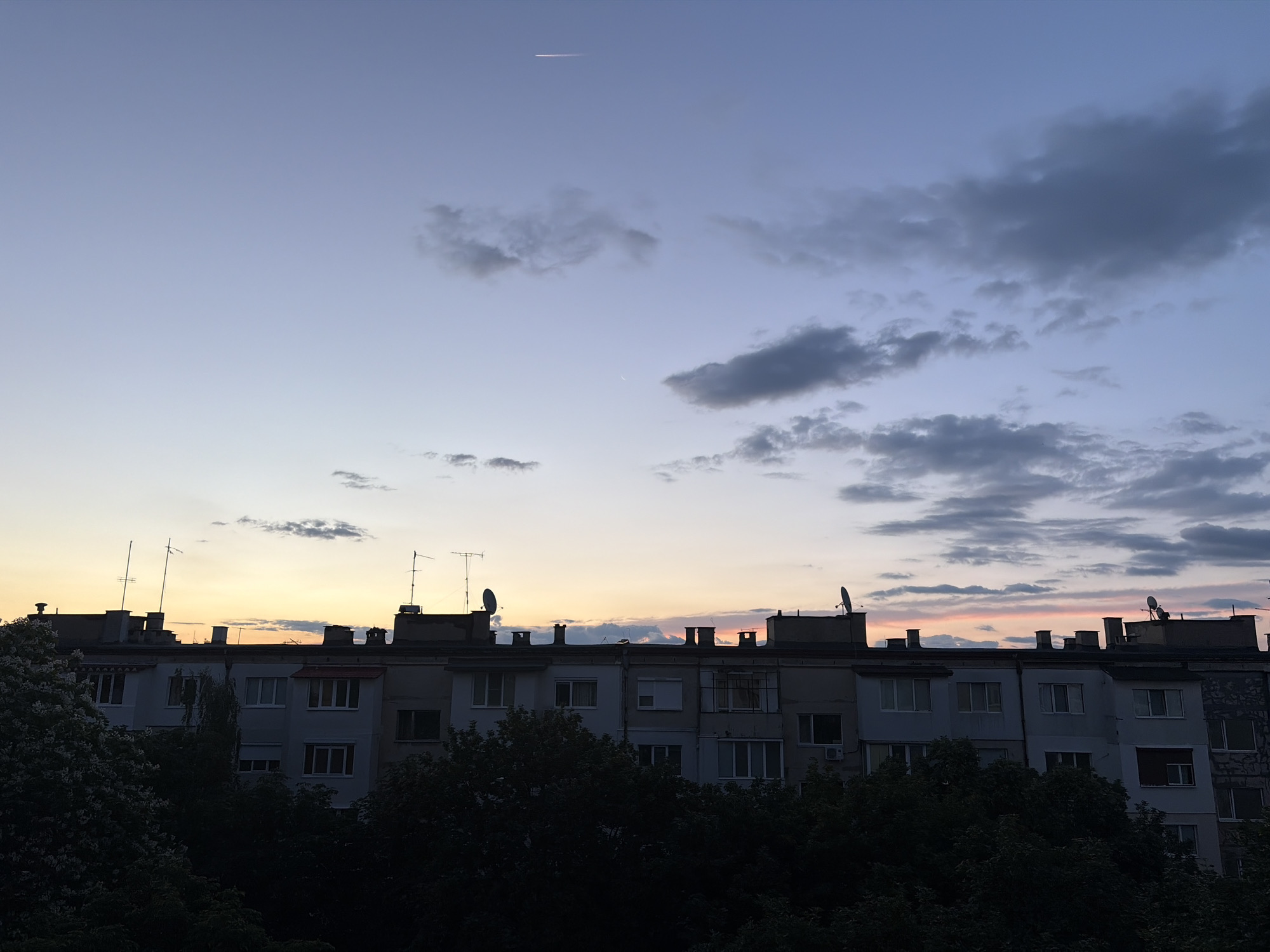 A wide shot captures a silhouetted apartment building against a twilight sky. The building, with multiple stories and numerous windows, runs horizontally across the frame, its dark form partially obscured by the dense, dark green foliage of trees in the foreground. The sky above transitions from a pale blue at the top to soft hues of orange and pink near the horizon, indicating either sunrise or sunset. Wispy clouds are scattered across the sky, with a few darker, more defined clouds present. Antennas and satellite dishes are visible on the rooftop of the apartment building. A faint contrail from an aircraft is visible high in the sky. The scene is static and devoid of any visible human activity.