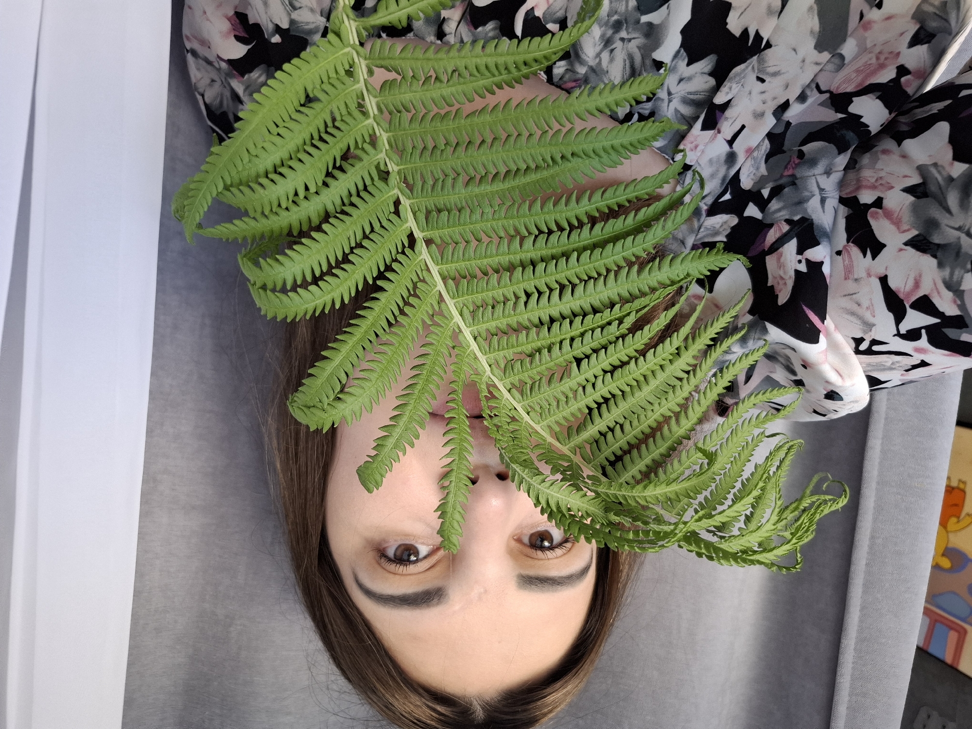 In a medium shot, a woman holds a large, green fern frond in front of her face, obscuring her nose and mouth. Her eyes, which are brown, are visible above the leaves. She has medium-length brown hair and is wearing a floral-patterned top with black, white, pink, and gray colors. The background consists of gray and white curtains. A small portion of a colorful artwork depicting a cartoon character can be seen on the far left of the image. The scene appears to be indoors, possibly in a residential setting in Chaikovsky, Russia.