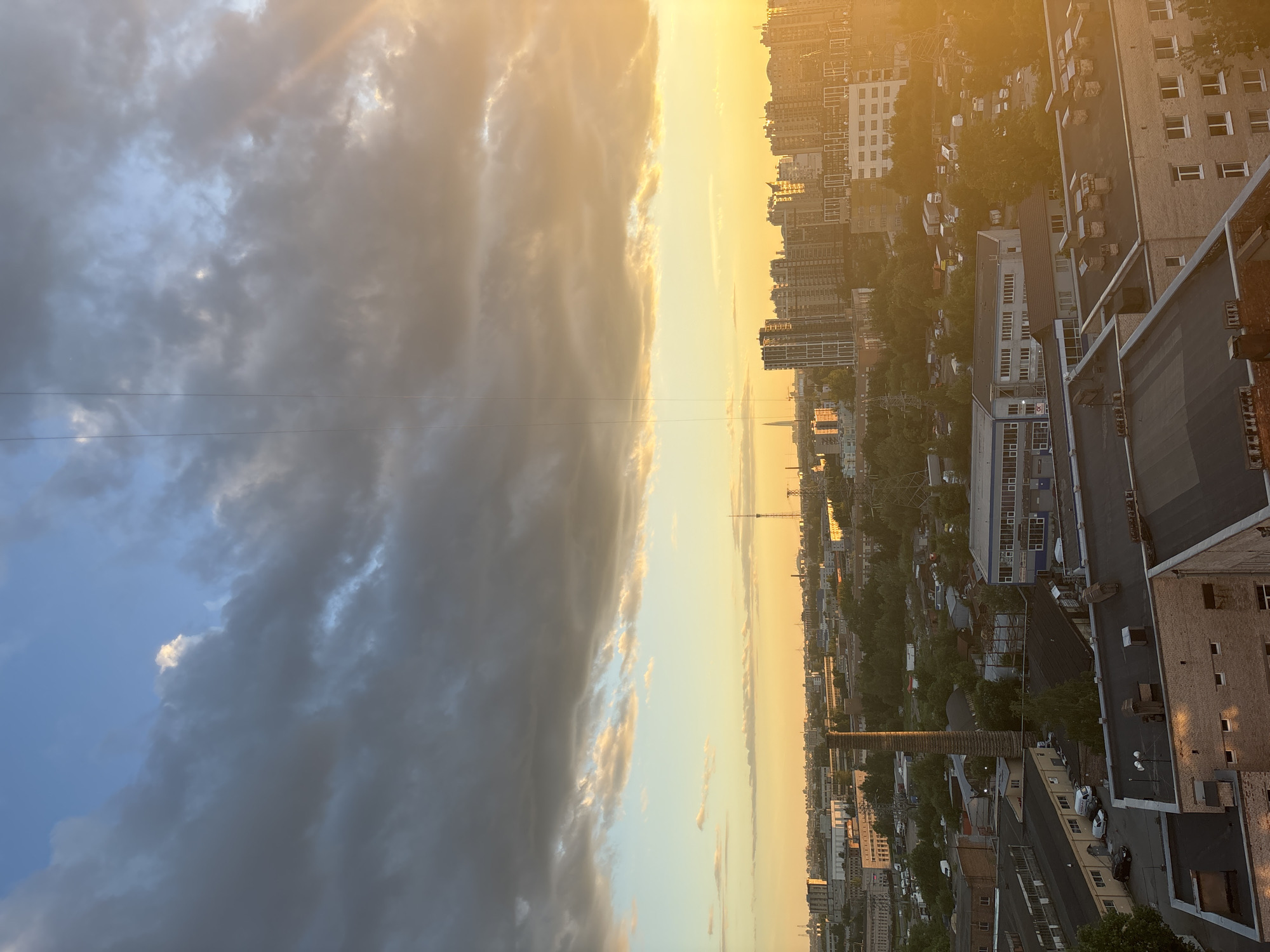 A wide shot captures an urban industrial landscape at sunset. Dark gray clouds dominate the upper half of the frame, with pockets of lighter, illuminated clouds and a bright yellow sunburst emanating from the upper right corner. The lower half of the image shows a densely built cityscape with a mix of residential and industrial buildings, characterized by flat roofs. A tall, cylindrical brick chimney stands prominently in the mid-ground. In the distance, a cluster of modern high-rise buildings and a prominent tower, likely the Lakhta Center, are visible, suggesting a location in Saint Petersburg, Russia. The overall lighting is warm and golden due to the setting sun. There are no people or discernible actions visible in the image.