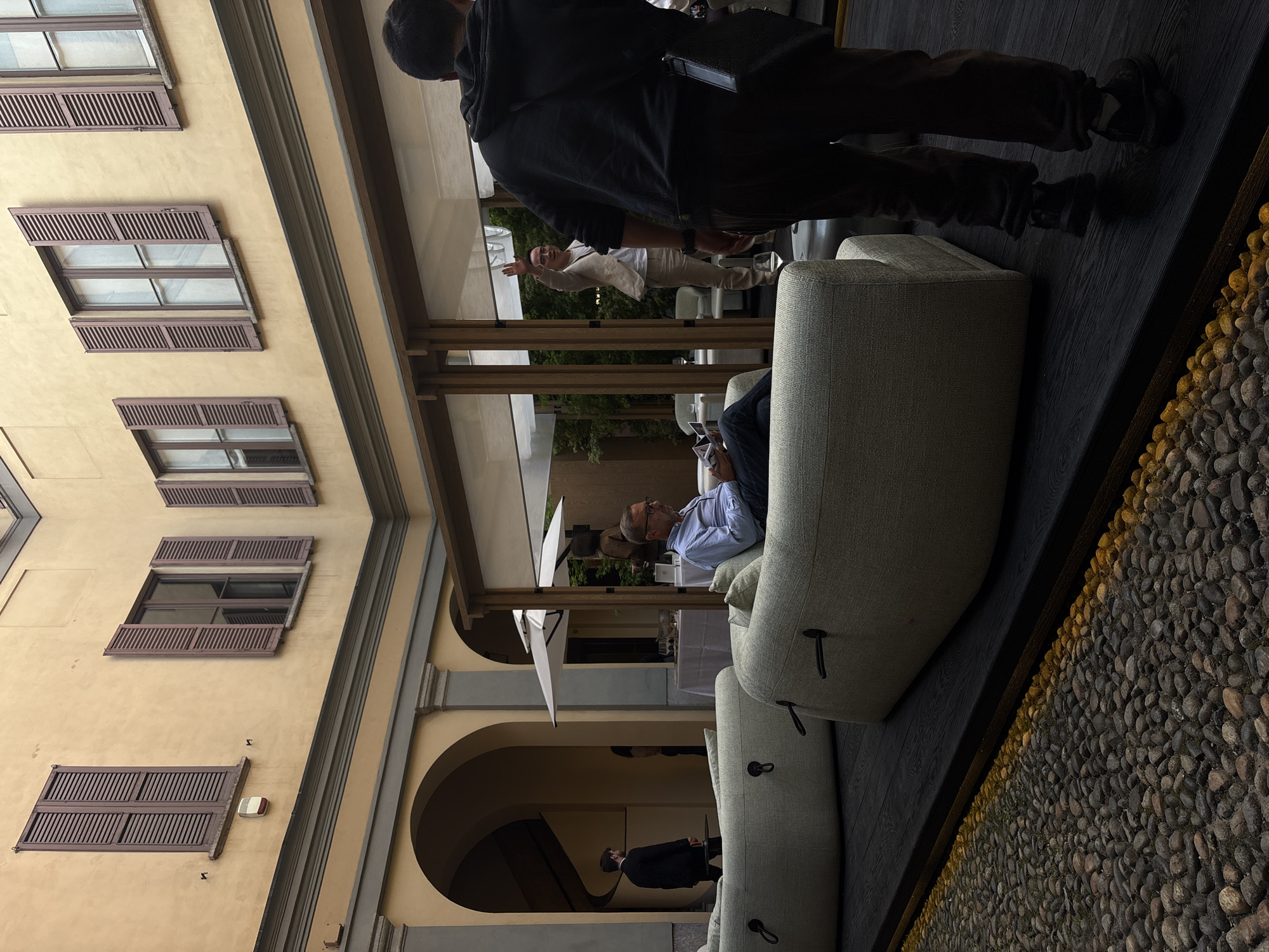 This image captures a vibrant scene within a sophisticated lounge or patio area, opening onto a historic courtyard, likely in Milan, Italy.
The setting features a warm, light yellow building facade on the left, characterized by multiple windows fitted with closed brown louvered shutters. Some windows are open, revealing glass panes. Arched passageways are visible on the lower level of the building, hinting at further architectural details. To the right, a dark wooden floor leads to a decorative border of small, dark, rounded pebbles, illuminated by a strip of golden-yellow light. Overhead, a structure with dark wooden beams and white awnings or blinds suggests a covered outdoor space.
Several individuals populate the scene. In the immediate foreground, with their back partially to the camera, a person wearing a dark hoodie and dark trousers stands, observing the area. Towards the center-right, a man with glasses, dressed in a light blue shirt and dark trousers, sits comfortably on a modern, light-colored modular sofa, engrossed in a device, possibly a phone or tablet. Behind this sofa, another man in a light-colored suit jacket and trousers stands with a visible smile, his arm raised in a welcoming or gesturing manner. Further back, beneath one of the building's arches, a person in a dark suit or jacket is discernible. A white table with a tablecloth and an umbrella are also visible in the background, suggesting a dining or refreshment area.
The overall atmosphere appears calm and relaxed, consistent with daytime leisure. The mix of classic Italian architecture with modern lounge furniture creates a blend of historical charm and contemporary comfort. No visible text is present in the image.