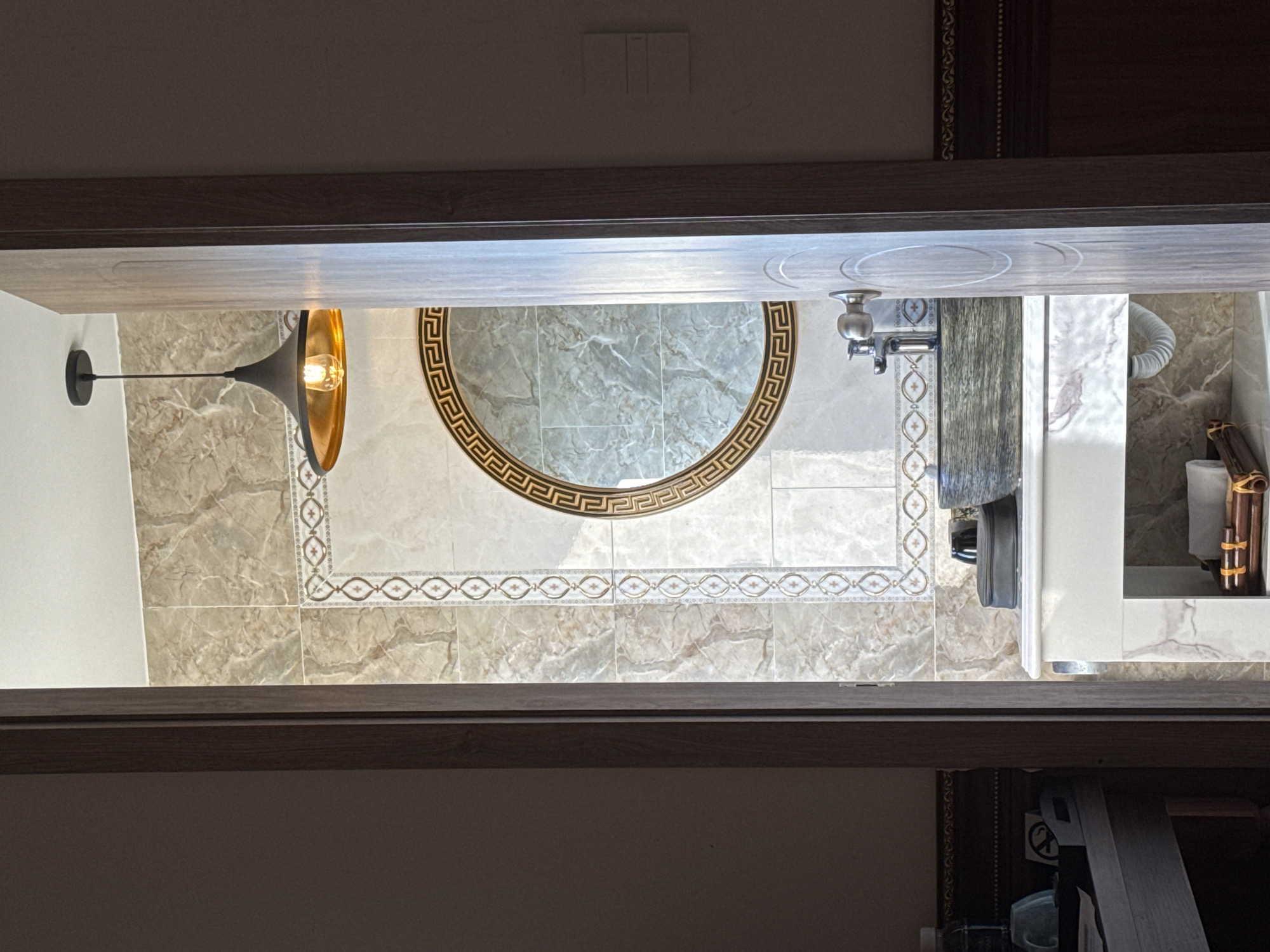 The image shows a top-down view of a bathroom interior. A round, decorative mirror hangs above a vanity, featuring a faucet. A light fixture is installed on the left wall. The floor is tiled with patterned marble. The image orientation is inverted. The location, Đà Nẵng, Vietnam, is not discernible from the image.