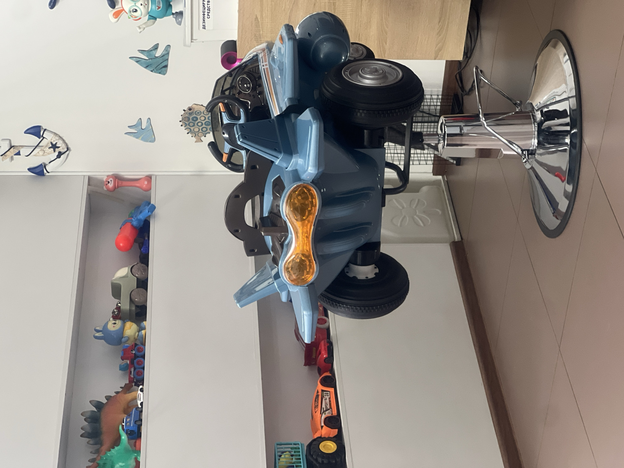 The image captures an interior scene, likely a children's salon or play area, in the city of Unknown, Unknown. Dominating the foreground is a child's styling chair designed as a blue and grey airplane-like vehicle, equipped with a steering wheel, dashboard, and orange-tinted headlights. This novelty chair is mounted on a chrome hydraulic base with a circular, reflective foot pedal, positioned on a light brown tiled floor with visible grout lines.
Along the left wall, three white shelves are filled with children's toys. The top shelf displays a green dinosaur, a brown dinosaur, a blue police car, a white toy car, a blue and red toy truck, a blue rabbit figurine, a light green pickup truck, a red and blue water gun, and a pink rattle. The bottom shelf holds a large yellow and blue construction truck, an orange sports car labeled "SPORTS 30," and two red toy cars.
The white wall on the right is decorated with a white and blue anchor, several light blue fish cutouts, and a patterned fish decoration. A white rabbit figurine wearing glasses and a blue shirt stands next to a white box bearing Russian text, "ДЕЗИНФИЦИРУЮЩИЕ СРЕДСТВА" (Disinfectants). A light wooden cabinet is partially visible on the far right. A dark brown baseboard runs along the bottom of the walls. The area is brightly lit, with no individuals present.