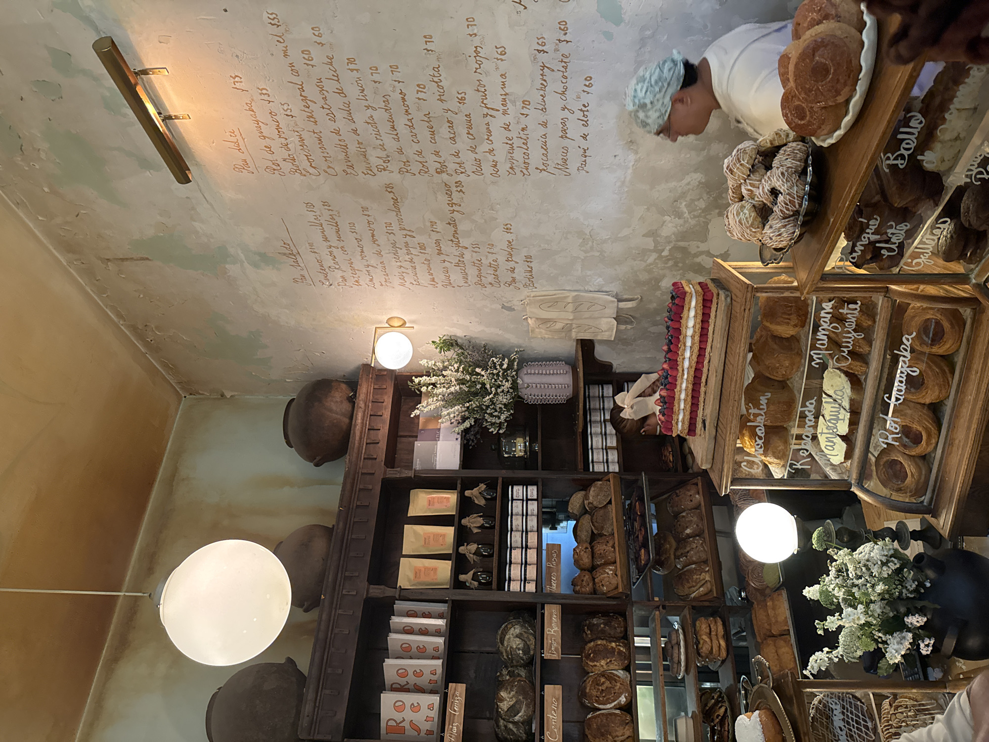 The image shows the interior of a bakery or pastry shop in Mexico City, Mexico. The shop features a variety of bread and pastries, including bolillos, baguettes, croissants, and other sweet and savory baked goods. The prices of the items are written on the wall. There are also shelves stocked with packaged goods. A person wearing a chef's hat is seen behind a display case of pastries. The lighting in the shop is warm and inviting, with globe-shaped lights hanging from the ceiling. The walls have a textured, rustic finish, and there are decorative items such as ceramic pots and floral arrangements.