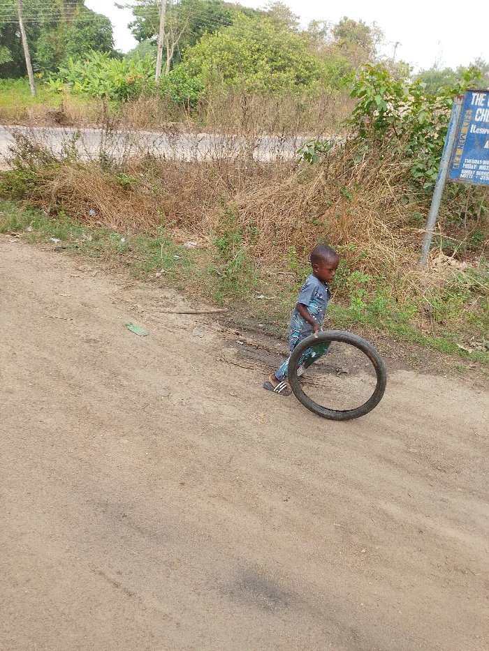 A young child is playing with an old tire on a dirt road. The child, dressed in patterned clothing and sandals, pushes the tire forward. The road runs along a grassy embankment. Behind the child is a growth of wild vegetation and some trees. There is also a signpost nearby. The sky is overcast.
