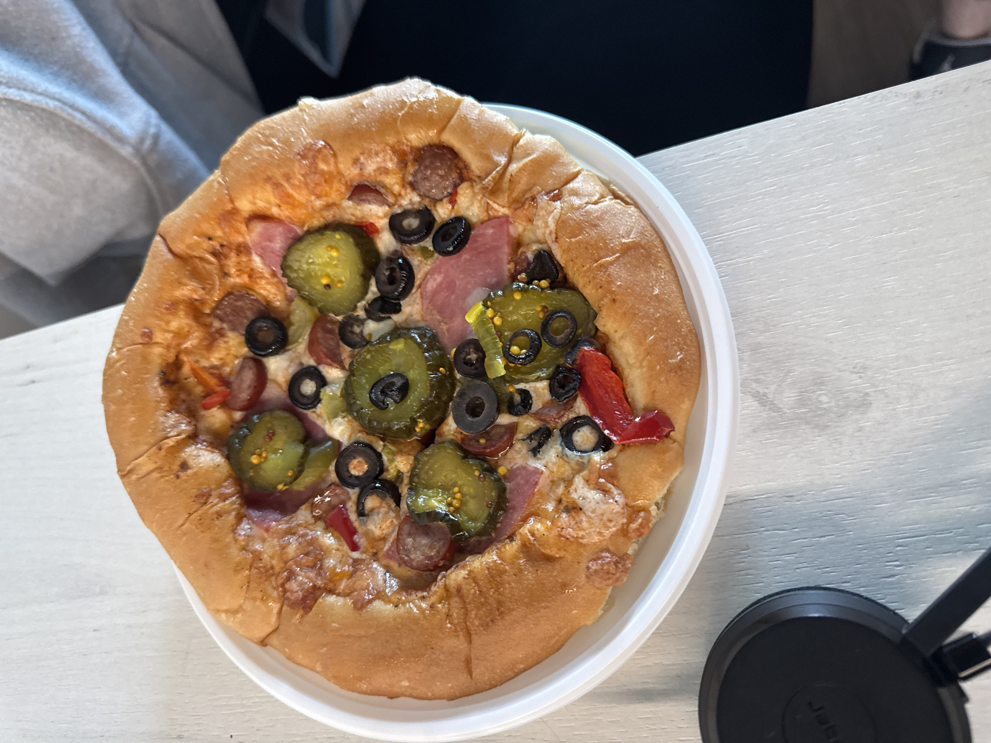 A close-up overhead shot shows a pizza on a white plate. The pizza has a thick, golden-brown crust, cheese, sliced olives, red peppers, sliced pickles, and pieces of sausage. The pizza is on a light-colored, textured surface that appears to be a wooden table. A black circular object with some writing on it is visible in the lower left corner of the image. The background is out of focus, with hints of a grey hooded sweatshirt. The setting is domestic.