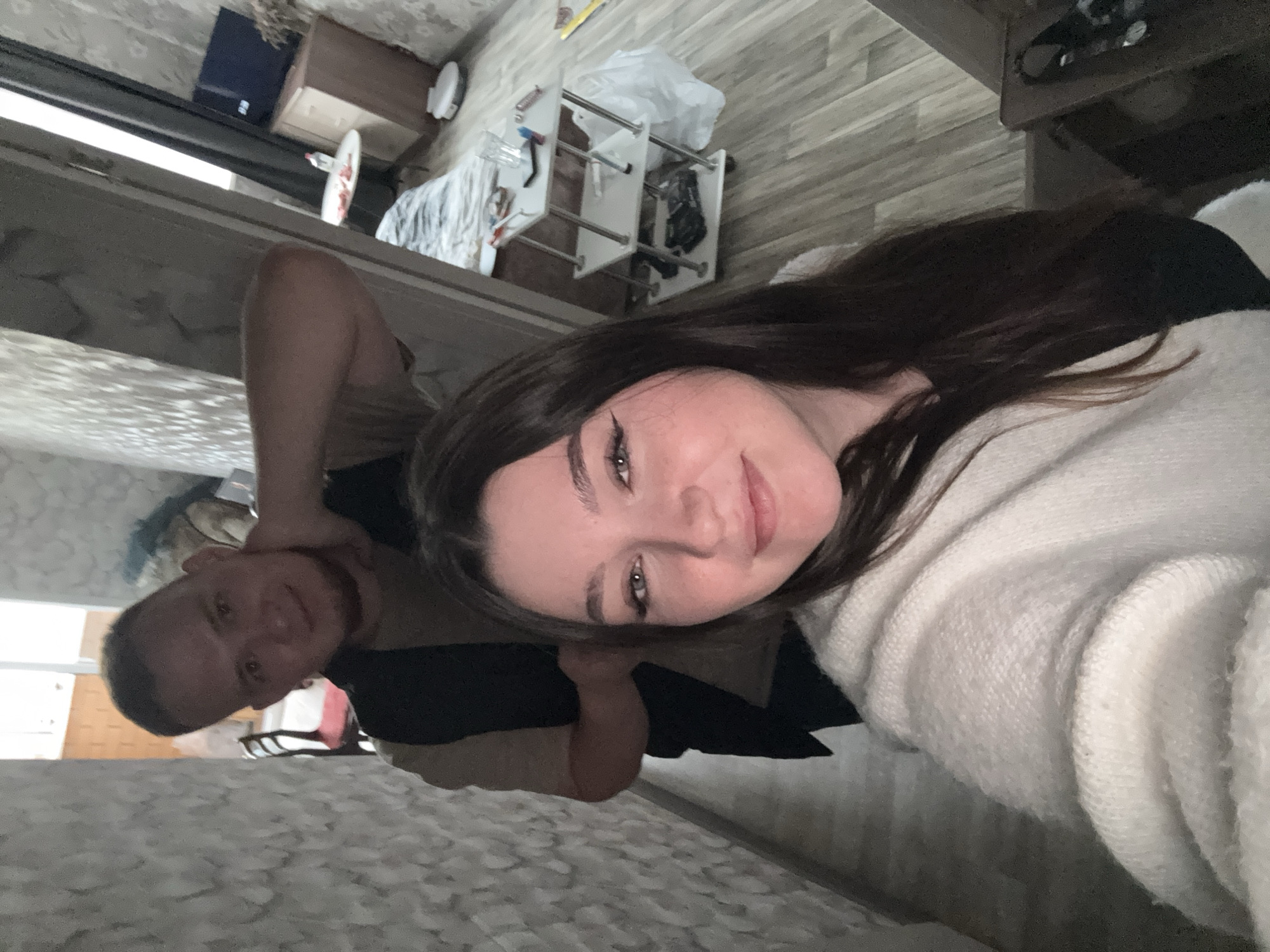 A selfie taken indoors in Perm, Russia. The photo features two individuals: a fair-skinned woman with dark brown hair, wearing a white sweater, and a fair-skinned man with short, dark hair. The man is slightly behind the woman, with his hand near his neck. The room is generally neat, with furniture and domestic items visible in the background, including a wooden cabinet, a metal rack with items, and a patterned wall. The floor appears to be covered with gray-toned wood-like material. The lighting is soft and diffused.