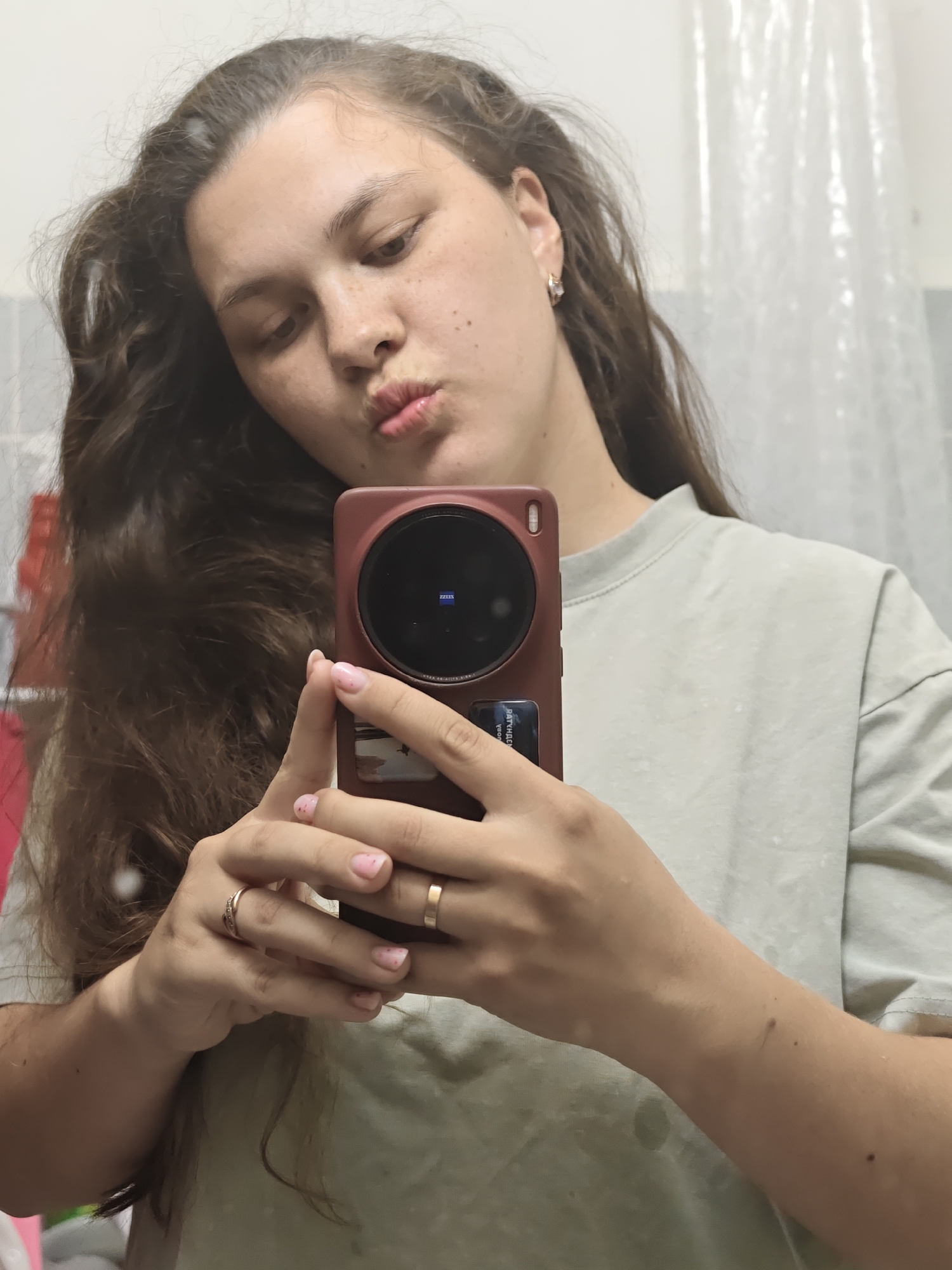 A woman takes a selfie in a bathroom. She has long brown hair, freckles, and is wearing a light gray t-shirt. She is holding a smartphone with a large circular camera lens, and her mouth is pursed as if posing for a kiss. She wears two rings on her left hand and a hoop earring on her left ear. The background shows a shower curtain and tiled walls, indicating a bathroom. The location in the city of Chaikovsky, Russia, is unknown.