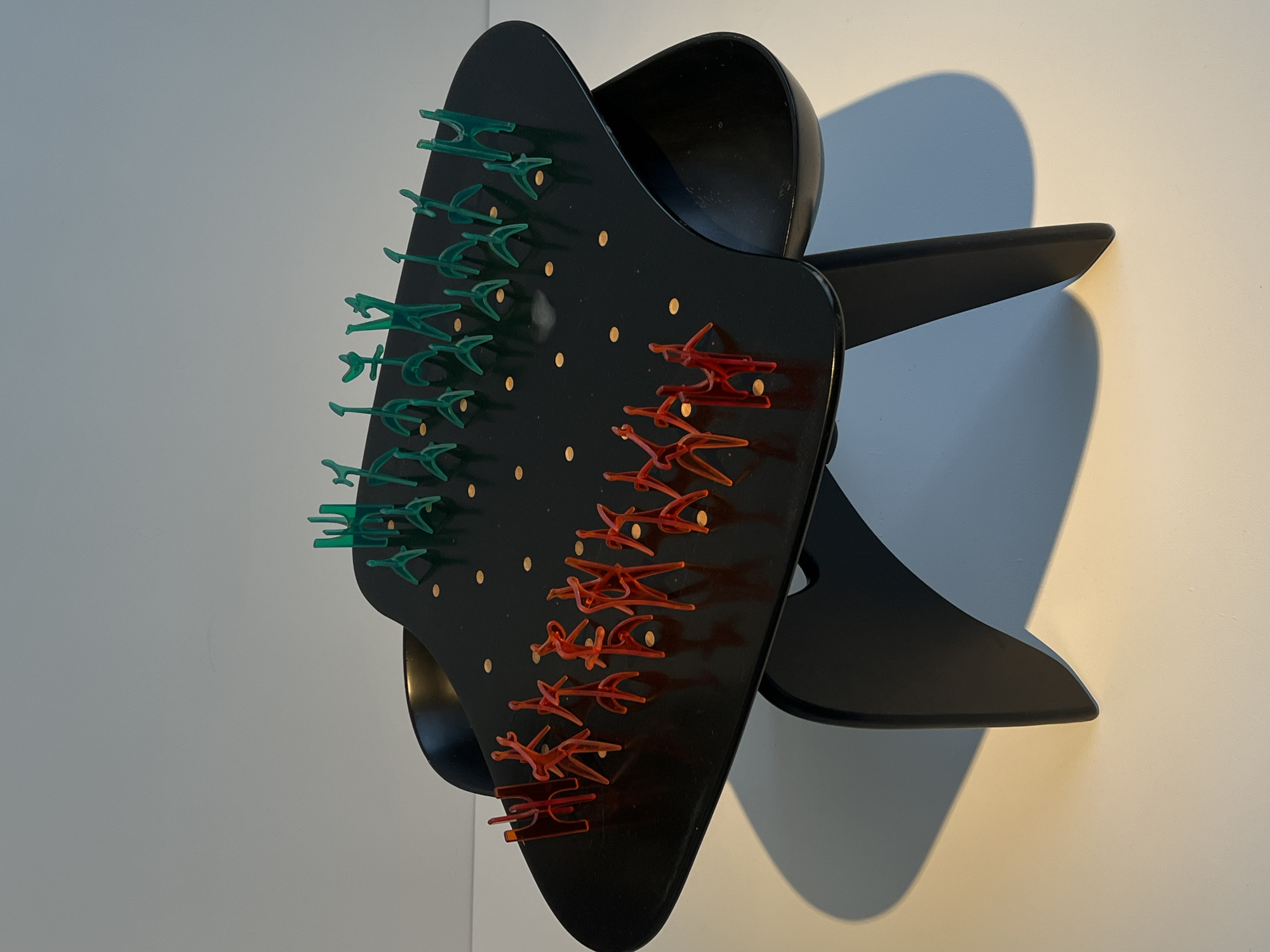 A close-up studio shot depicts a black, three-dimensional abstract object mounted on a light-colored wall. The object features a flat, black, roughly triangular plate with rows of green and red plastic shapes attached to it. These shapes appear to be stylized representations of vehicles or aircraft. The plate also has small, evenly spaced golden dots. Behind the plate, there's a black, curved structure that adds depth to the object. The object is lit from the side, casting a distinct shadow on the wall behind it. The image does not provide sufficient information to determine its location in New York City or provide any information about specific people, interactions, or actions. The scene is focused solely on the object itself.