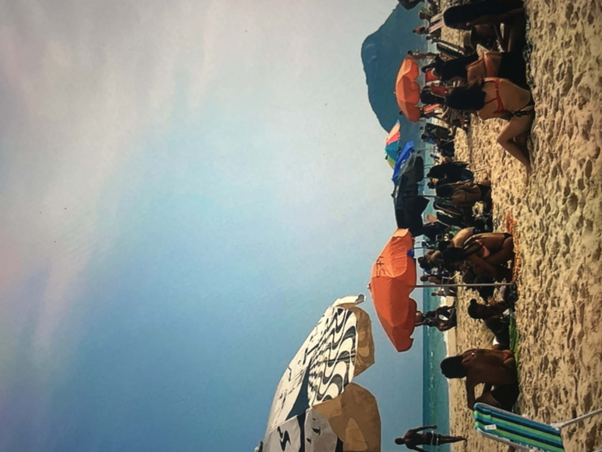 A group of people are enjoying a sunny day at the beach. Several colorful umbrellas provide shade on the sandy shore, with beach chairs scattered around. In the background, a calm ocean meets a clear blue sky, with a distant mountain range adding to the scenic view. Some individuals are sitting on towels, while others are walking along the water's edge. The atmosphere appears relaxed and festive, with many people dressed in swimwear.