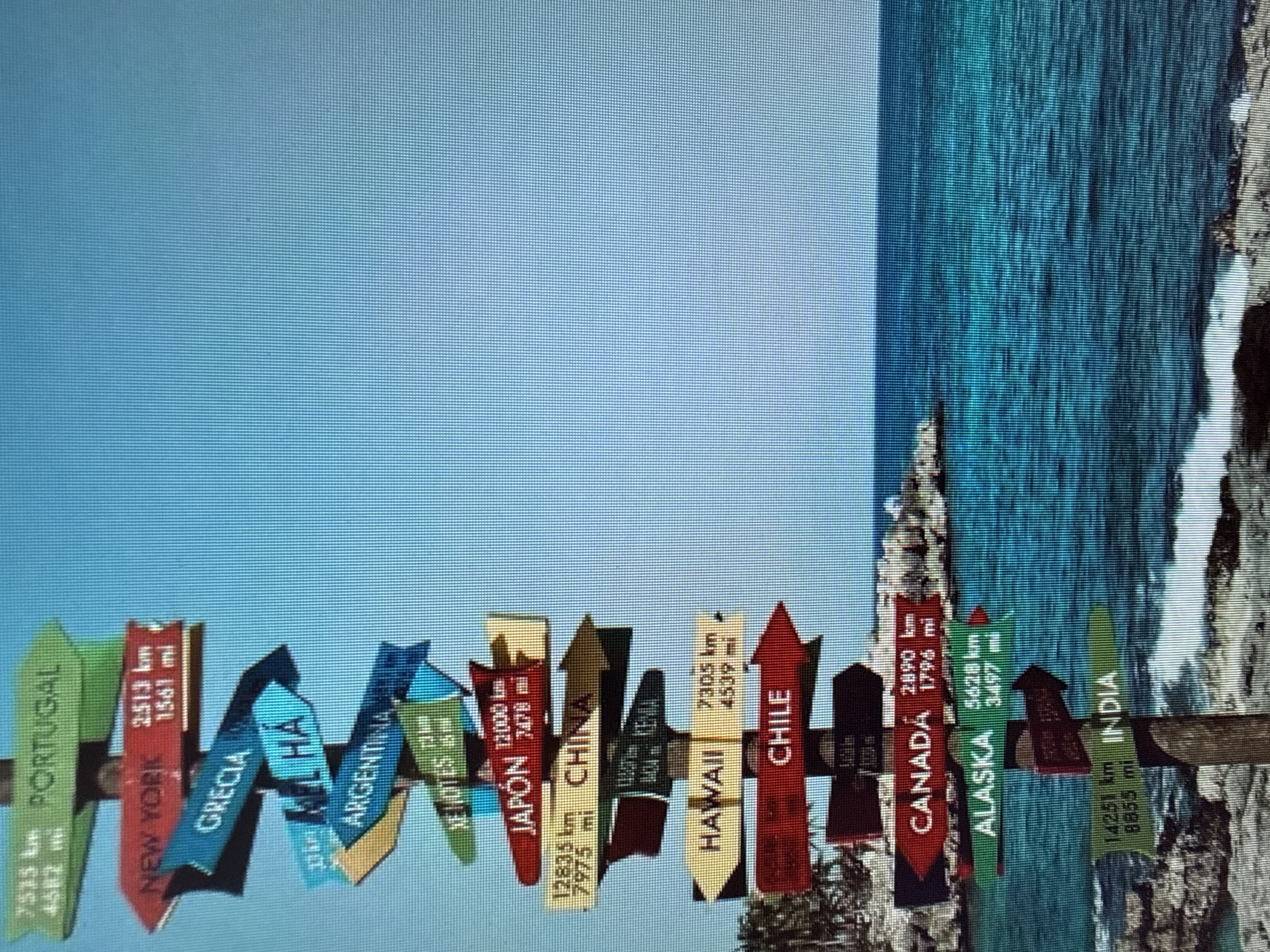 This image depicts a signpost with multiple directional arrows pointing to various international destinations. The signpost is made of wood and is weathered. Each arrow is colored differently and displays a destination name along with its distance in kilometers and miles.
The destinations listed are:
* Portugal
* New York
* Grecia
* El Hâ (likely a misspelling of El Hasa or similar)
* Argentina
* Xe Hotes (likely a misspelling of exotic hotels or similar)
* Japón
* China
* Kenya
* Hawaii
* Chile
* Canada
* Alaska
* India
The signpost is set against a backdrop of a clear blue sky and a turquoise ocean with a rocky coastline and some vegetation visible in the distance. The overall impression is one of travel and exotic locations. The location is not specified to be within a city, but rather in a natural setting conducive to tourism. There are no people or specific actions depicted in the image.