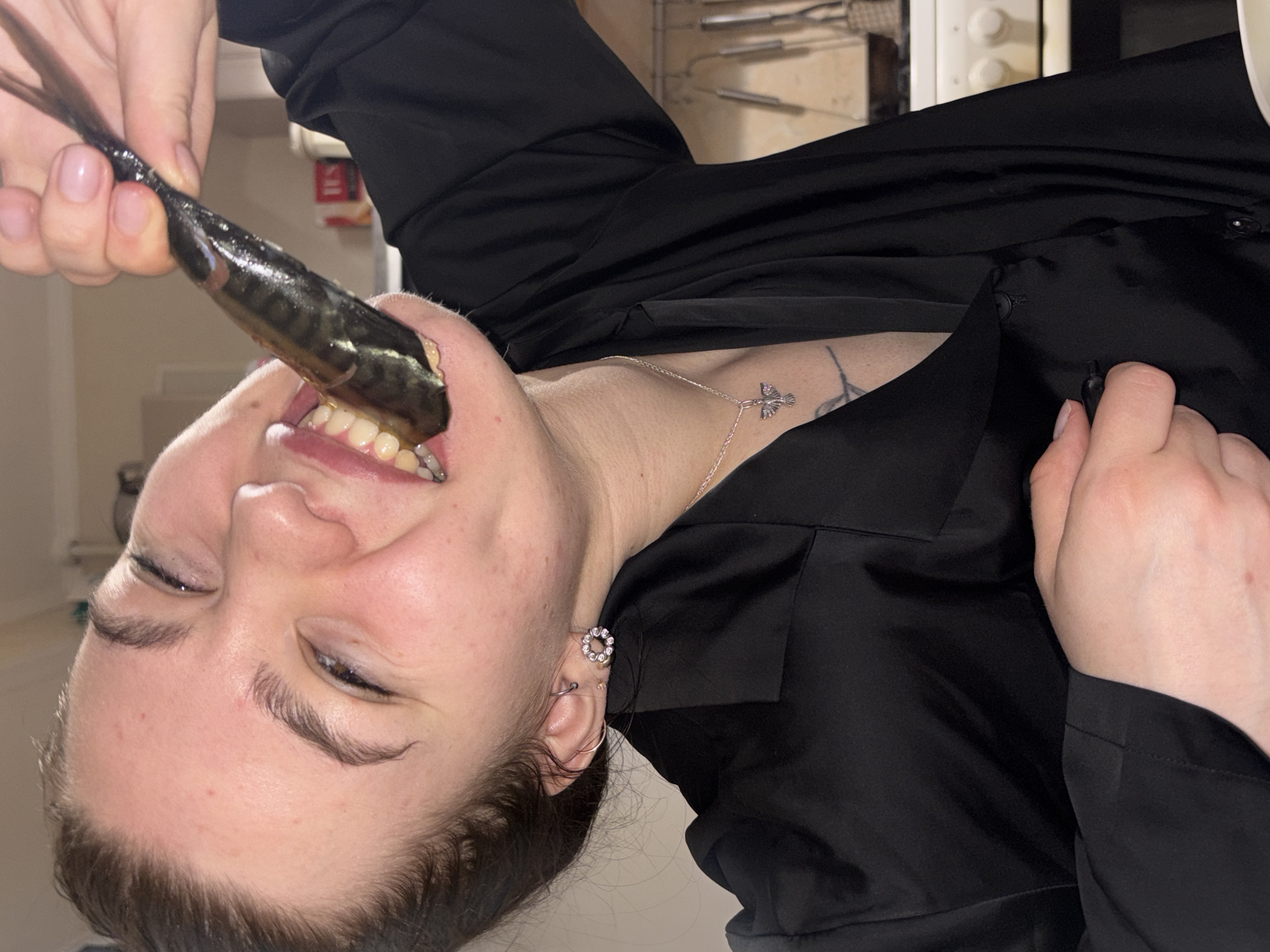 A woman is holding a whole fish, possibly a mackerel, with her hand and inserting it into her mouth. She is wearing a black, shiny collared shirt, a silver necklace, and an earring. She also has a tattoo on her neck. The location in Perm, Russia, cannot be determined from the image.