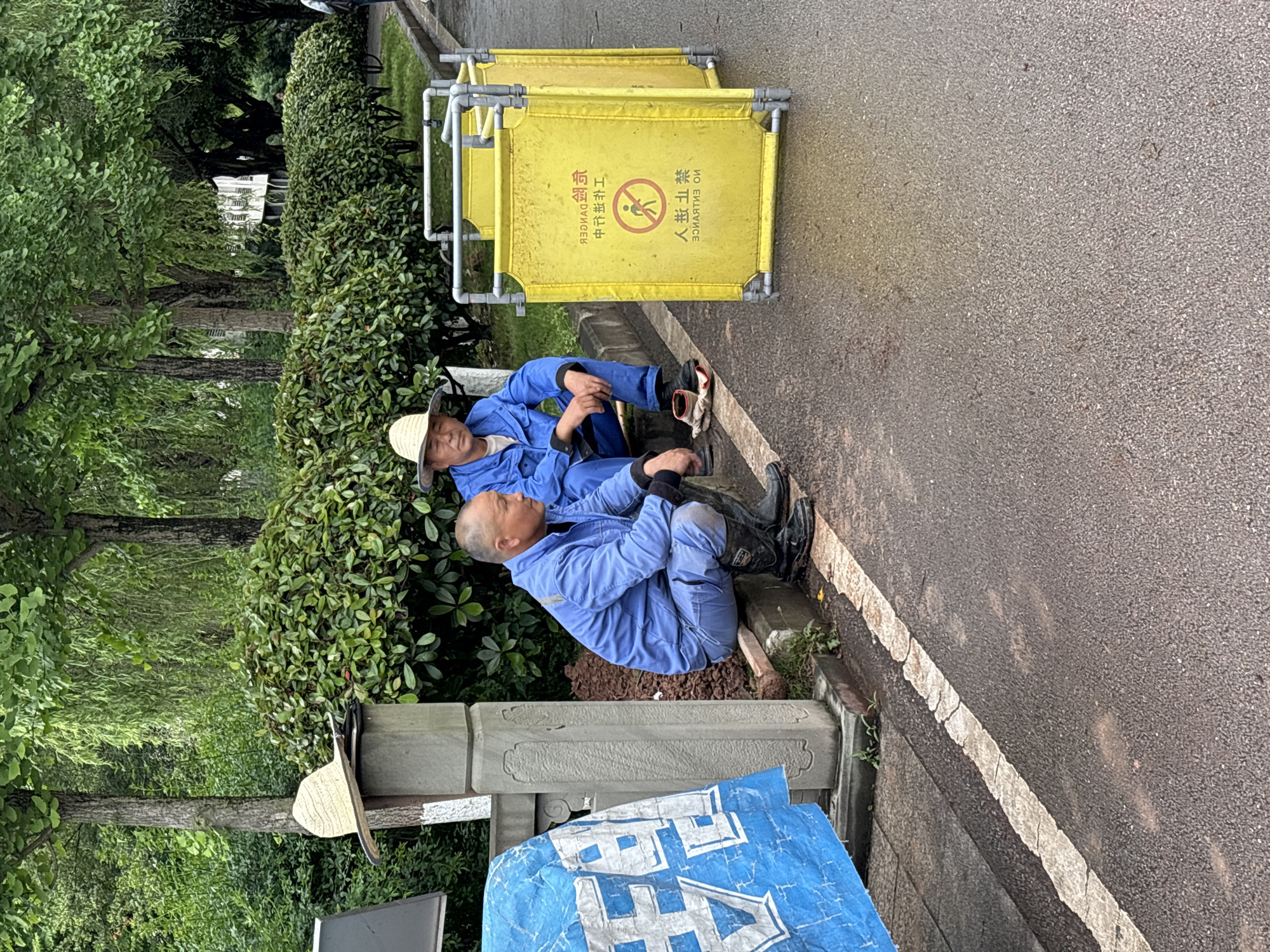 Two men in blue work uniforms are resting by the side of an asphalt path in Jiangjin, China, amidst lush greenery. One man, wearing a straw hat, sits with his back against a dense green bush. Further down, another man, also in a blue uniform, sits in a similar posture, leaning against the foliage.
Next to them are yellow foldable safety barriers, typical for construction or maintenance areas. The ground is asphalt with a faded white line, appearing damp, suggesting recent rain or high humidity. The path is bordered by thick green bushes and taller trees in the background. A decorative stone pillar with carved details stands near the foreground. A blue tarp or fabric with large white Chinese characters is also visible in the lower left.
The yellow barriers display Chinese text: "注意安全" (Zhùyì ānquán), meaning "Pay attention to safety," and "人行禁止" (Rénxíng jìnzhǐ), meaning "No pedestrian access," accompanied by a universal "no entry" pictogram (a person within a red circle with a diagonal line). Reversed English text "NO ENTRANCE" is also faintly visible on the barrier. The large white characters on the blue tarp appear to spell "禁止" (Jìnzhǐ), meaning "Forbidden" or "Prohibited."
The scene captures a moment of rest for the workers during the daytime, likely in an outdoor setting such as a park or roadside.
