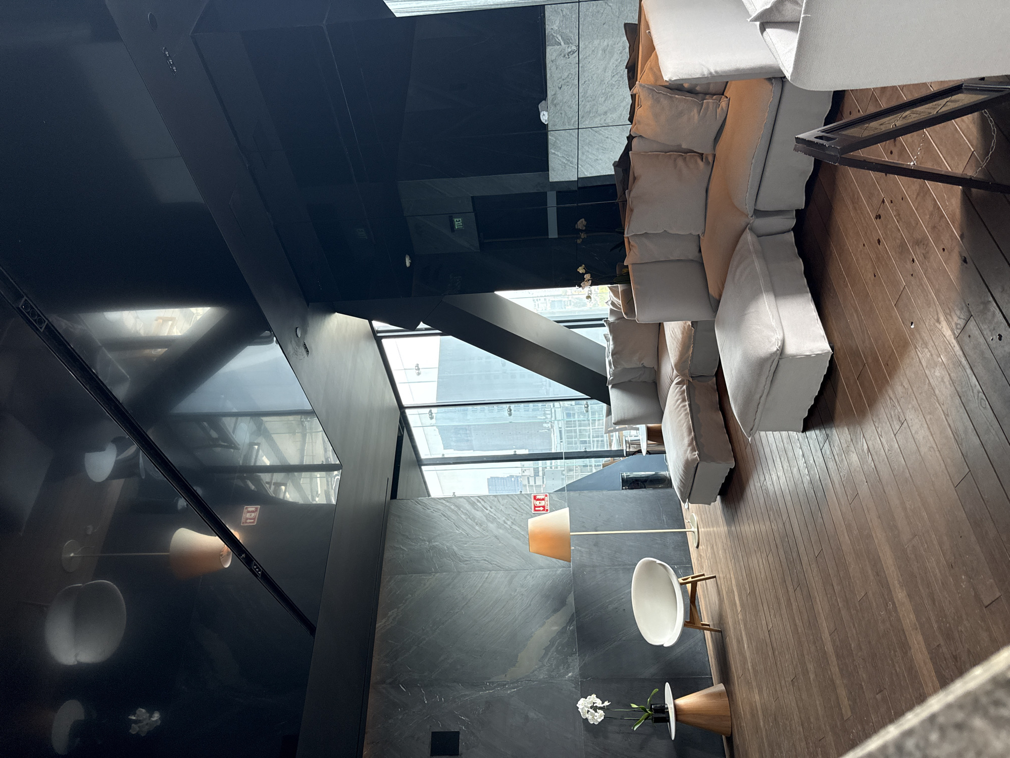 The image shows an interior view of a modern-designed room, possibly in Mexico City, Mexico, suggested by the architectural style and interior design choices. The room features large windows offering an urban view, indicative of a city environment.
The room has a minimalist and luxurious aesthetic, with a dark ceiling and a light-colored sectional sofa with numerous cushions dominating one side. The floor is made of wooden planks, and a small table with a white chair sits nearby. A floor lamp with a beige shade and a small vase with white flowers add to the room's decor. The overall lighting seems to be a mix of natural light from the windows and artificial lighting from the lamps, contributing to a comfortable and stylish atmosphere. There are no people visible in the image.
