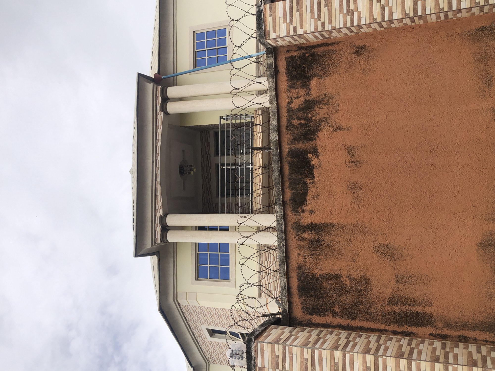 A low-angle, medium shot captures the facade of a two-story house in Unknown, Unknown, against a cloudy sky. The house features a light yellow exterior with a detailed trim along the roofline. Two large, fluted columns support a small balcony on the upper floor, which has a black metal railing. Blue-framed windows are visible on both floors. The house is surrounded by a fence; the lower part is a solid, textured reddish-brown wall, while the upper part is a section of brickwork. On top of the brickwork, barbed wire is stretched in a crisscross pattern. A long, blue pole with a red top leans against the house on the right side, extending from the balcony area upwards. The overall scene suggests a residential property with security measures.