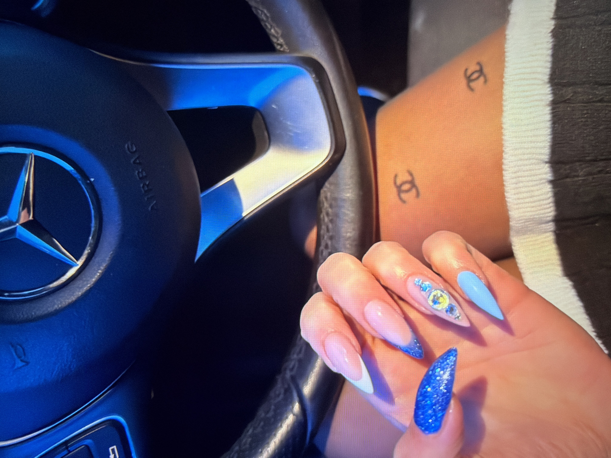 The image offers a close-up view inside a car, focusing on a person's hand, leg, and the steering wheel. A right hand, adorned with long, pointed nails, rests near a Mercedes-Benz steering wheel. The nails showcase diverse designs: the thumb is a solid glittery blue, the index finger features a blue glitter French tip, the middle finger has a nude base embellished with blue and yellow gemstones, the ring finger is a solid light blue, and the pinky has a classic white French tip.
The black steering wheel, prominently displaying the Mercedes-Benz star logo, features blue-tinted metallic accents. The word "AIRBAG" is visible on the upper part of the steering wheel. In the background, on what appears to be the person's upper thigh or leg, two small, black tattoos of the Chanel logo (interlocking C's) are visible. A section of white ribbed fabric, likely clothing or car interior trim, is present on the right side of the frame. The lighting suggests an indoor setting or nighttime, with a distinct blue glow highlighting components of the car's interior.