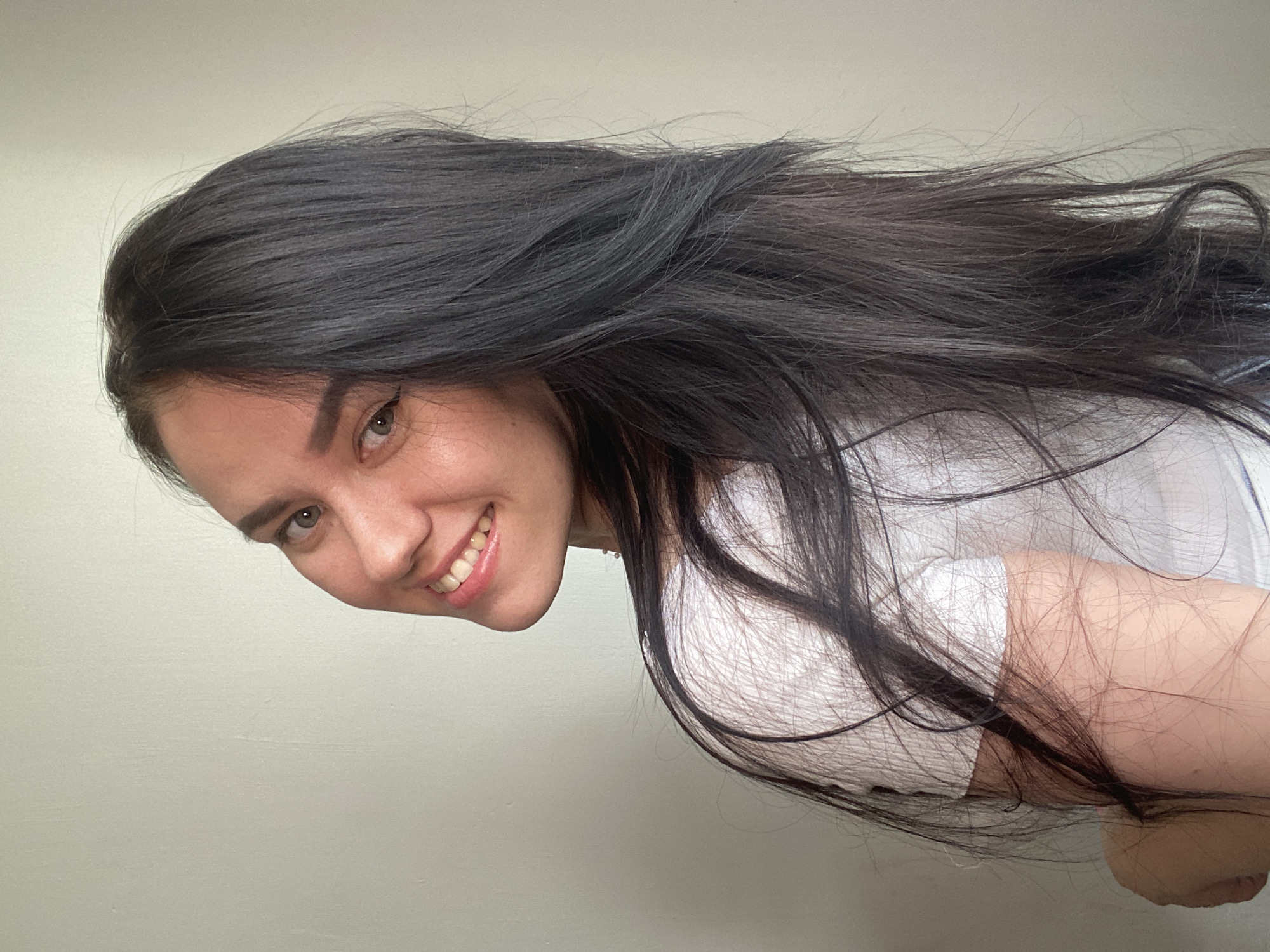 A young woman with long, dark hair is pictured in a close-up shot. She is looking over her left shoulder towards the camera, smiling broadly. Her hair is styled down and covers part of her right shoulder. She is wearing a white t-shirt. The background is a plain, light-colored wall. The location is not specified in the image.