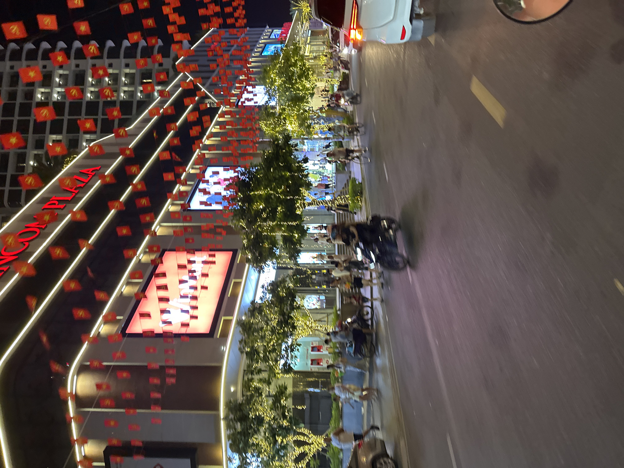 The image depicts a street scene at night in Nha Trang, Vietnam, likely on a festive occasion, given the abundance of red flags with yellow stars, resembling the Vietnamese flag, strung across the street and adorning buildings. A large building with illuminated signage, reading "NGCON PLAZA," is prominently featured. The plaza's facade is decorated with strings of small red and yellow flags and illuminated by modern architectural lighting. Trees lining the sidewalk are decorated with numerous small, bright yellow lights. People are visible walking on the sidewalk and crossing the street, while several motorcycles and a car are in motion on the road. The overall atmosphere suggests a lively urban environment, possibly a commercial or entertainment district. The foreground of the image shows a portion of a motorcycle's rear-view mirror and the street surface, indicating the photo was taken from a moving vehicle.