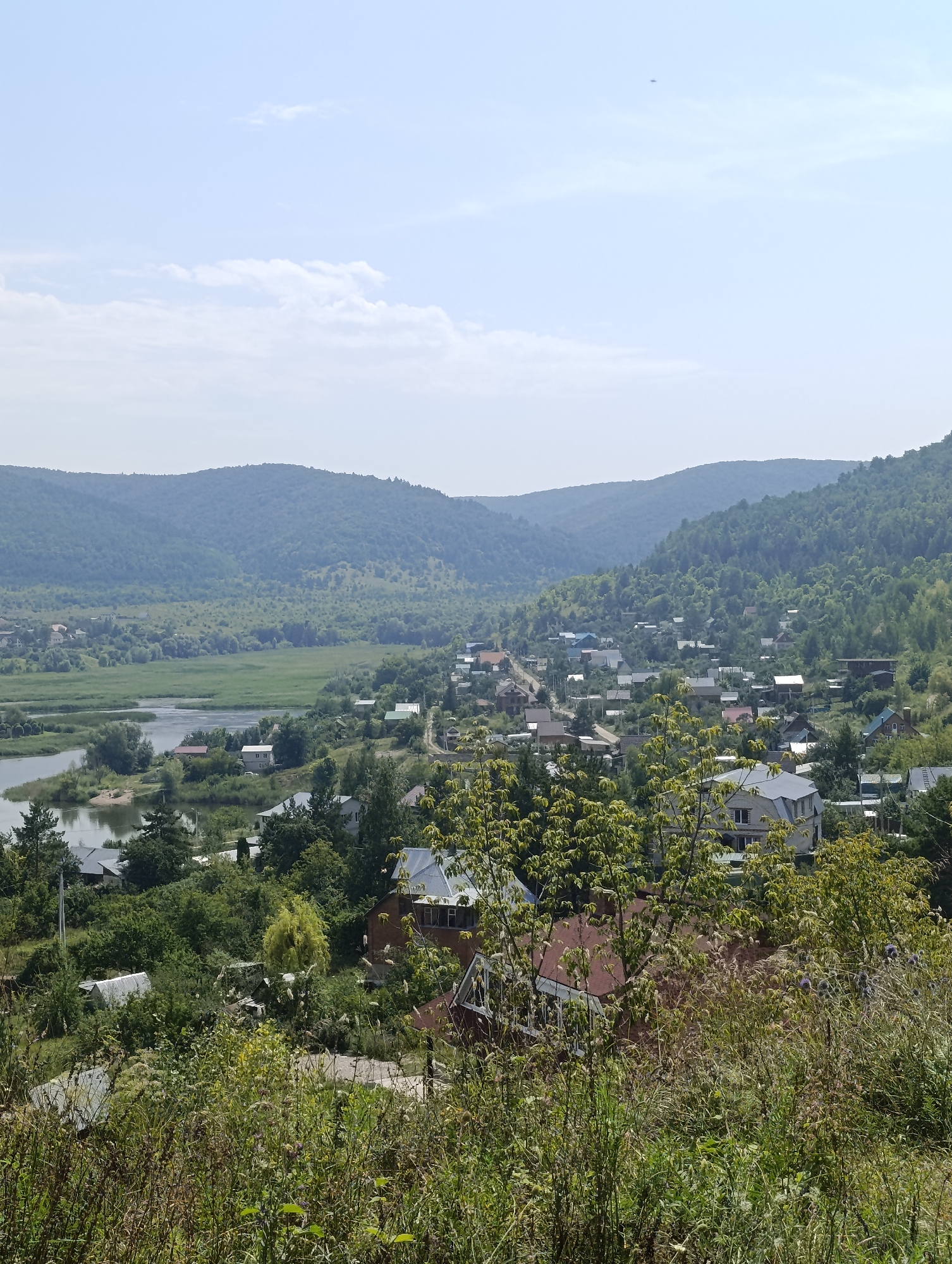 The image shows a panoramic view of the settlement of Solnechnaya Polyana, Russia, on a bright, hazy day. The foreground is dominated by a dense growth of dry grass and green foliage, obscuring the lower part of the scene. Beyond this, a village is nestled among rolling, tree-covered hills. Houses with pitched roofs, varying in color and architectural style, are scattered throughout the landscape. A river or large body of water winds its way through the valley, reflecting the sky and surrounding greenery. In the mid-ground, a lush green field stretches towards the water, bordered by trees. The background consists of several layers of receding, densely forested hills that fade into the hazy horizon. The sky is a pale blue with scattered, wispy white clouds. A small, dark object, possibly an aircraft, is visible high in the sky. The overall impression is of a tranquil, rural settlement set within a verdant natural environment.