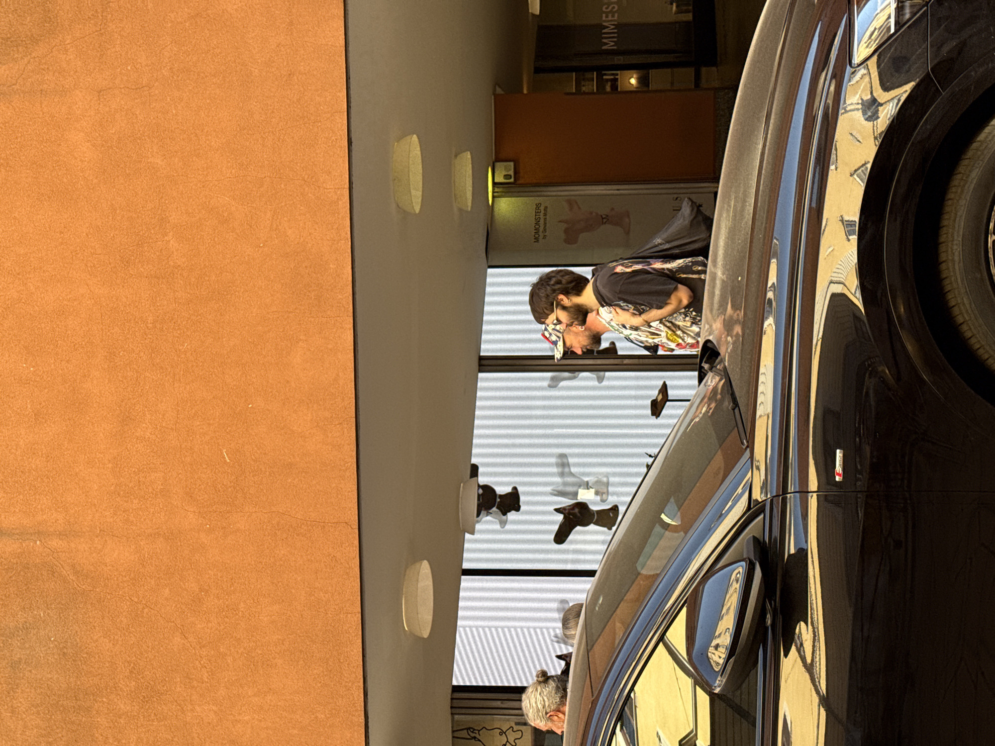 The image captures an outdoor scene in Milan, Italy. On the left is a textured orange wall adjacent to a neutral-colored building facade, marked with recessed lighting fixtures. A glass display window exhibits art pieces resembling limbs or stylized figures hanging in a row.
Two people are visible in the reflection of the window. One individual is wearing a cap and a printed shirt, while another behind them has dark hair and a beard. They appear to be looking at the exhibition.
The right side of the image features a black car with its door reflecting the surrounding buildings. A logo, likely a vehicle trim designation, is affixed to the side. The car's tire and wheel are prominently displayed on the right edge of the frame.