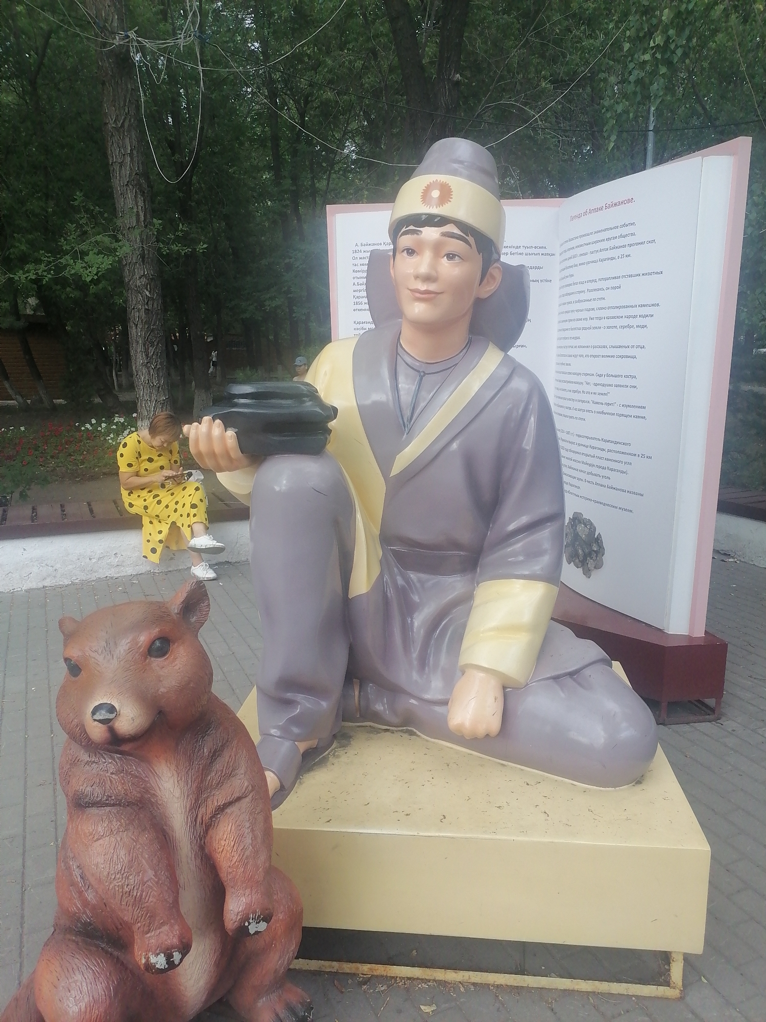 A statue of a young man in traditional attire sits on a large, light-colored pedestal next to an open book sculpture. The statue depicts the man with dark hair, a distinctive hat adorned with a floral emblem, and a flowing grey robe with yellow accents. He is holding a dark, bowl-like object in his right hand. In front of the statue, to the left, is a sculpture of a brown marmot sitting upright. Behind the main statue, an open book is displayed, featuring text in a foreign language. To the left of the main subjects, a woman in a yellow polka-dot dress is seated on a bench, looking down at her phone. She is wearing white sneakers. The environment appears to be an outdoor public space, with trees and foliage visible in the background, and paved ground in the foreground. The presence of statues and a public seating area suggests a park or a monument site within the city of Qarağandı, Kazakhstan.