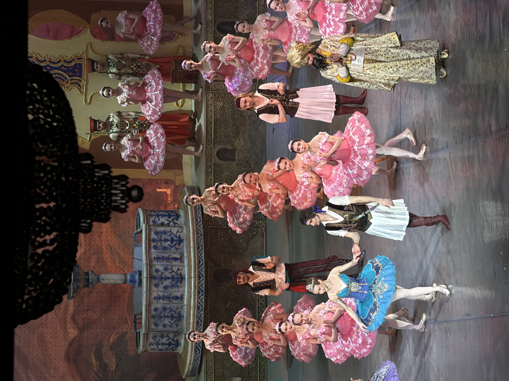 The image shows a scene from a performance, likely a ballet, taking place on a stage with a backdrop depicting architectural details. In the foreground, a group of ballet dancers are arrayed in various costumes, predominantly pink tutus for the female dancers, and some male dancers in what appear to be character costumes. The staging is intricate, with a tiered fountain or decorative structure visible on the left. Based on available details, this performance could potentially be taking place in Izhevsk, Russia, but this location cannot be definitively confirmed from the image alone. There are other stage and performance objects, such as lights, and other set pieces.
