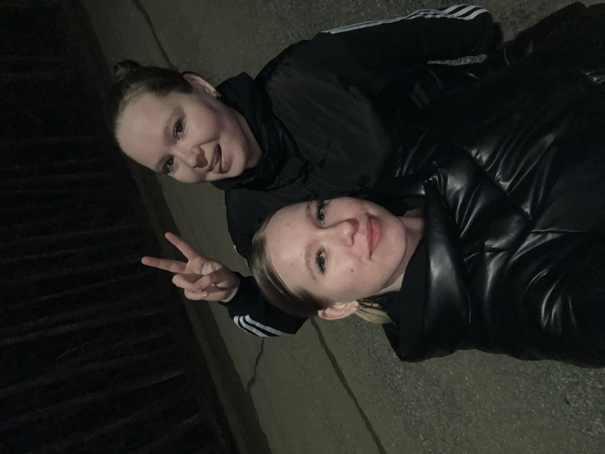 Two women are on the ground outdoors at night. One is lying on her back, wearing a black puffy jacket. The other woman is partially on top of her, wearing a black Adidas jacket with white stripes on the sleeves. She is making a peace sign with her fingers. The location is not specified. The ground is concrete.