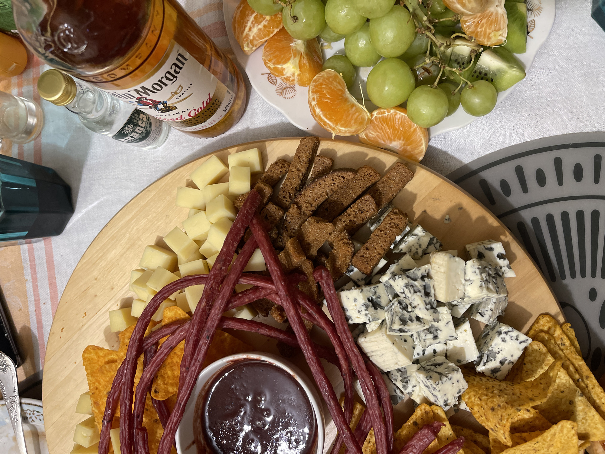 The image depicts a charcuterie board with various foods, including cubed cheese, long, thin meat sticks, small pieces of dark bread, blue cheese, tortilla chips, and a white bowl containing a dark-colored dip or sauce. Beside the board is a plate with grapes, tangerine slices, and kiwi. Two bottles, one of Captain Morgan rum and a smaller bottle of clear liquid, are also present. The scene is set on a surface covered with a white tablecloth. The image does not provide information to confirm the location is Ternopil, Ukraine.