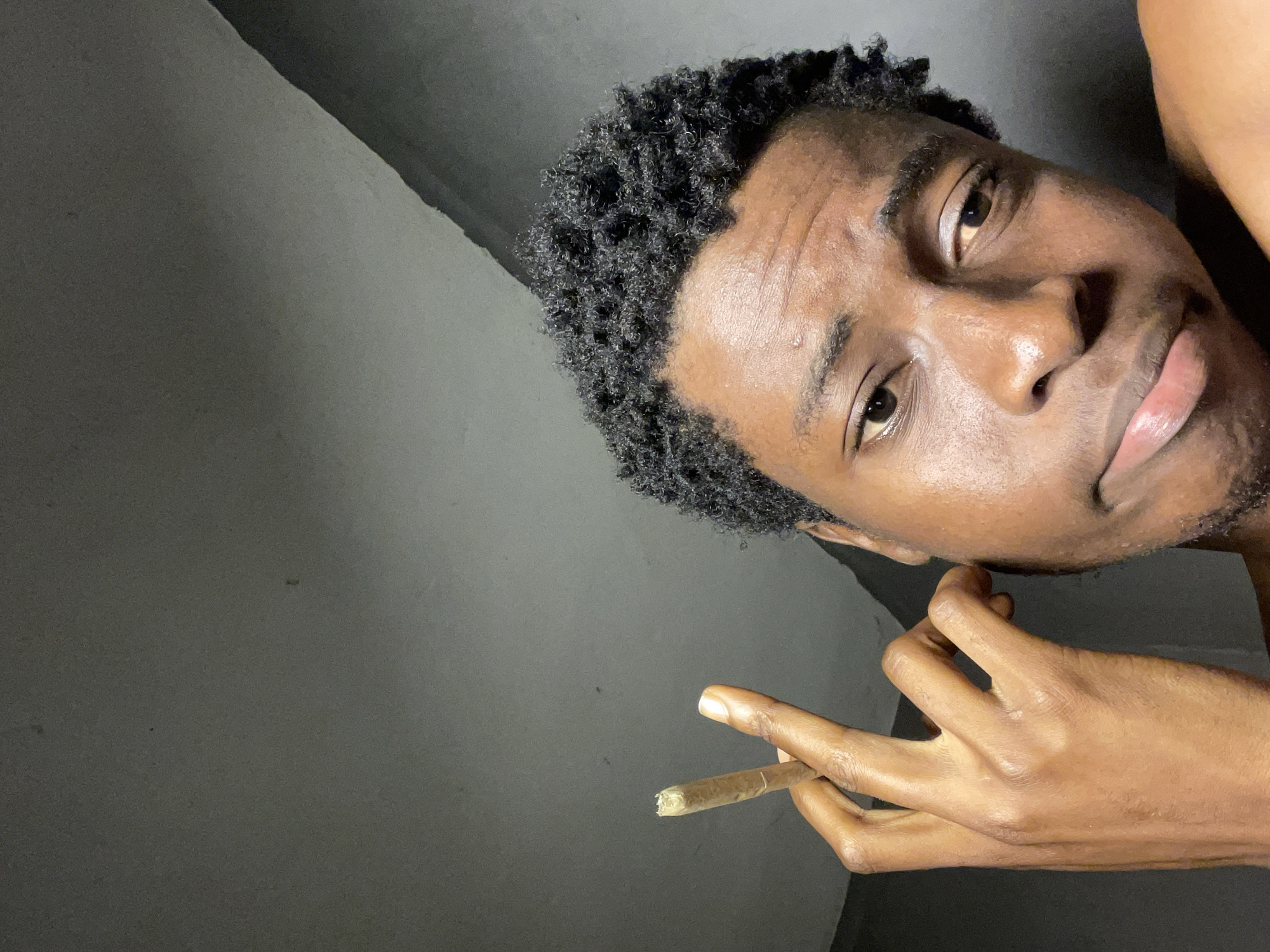 The image shows a close-up of a young Black person's face and hand. The person has dark, curly hair and is holding a rolled cigarette or blunt between their index and middle fingers. Their facial expression is neutral, with their eyes looking slightly to the side. The background appears to be a dimly lit, textured wall, possibly indoors. There is no indication of the specific location within Nkpor, Nigeria. The lighting suggests it may be evening or a room with artificial light. The overall scene is intimate and personal, focusing on the subject and their action.