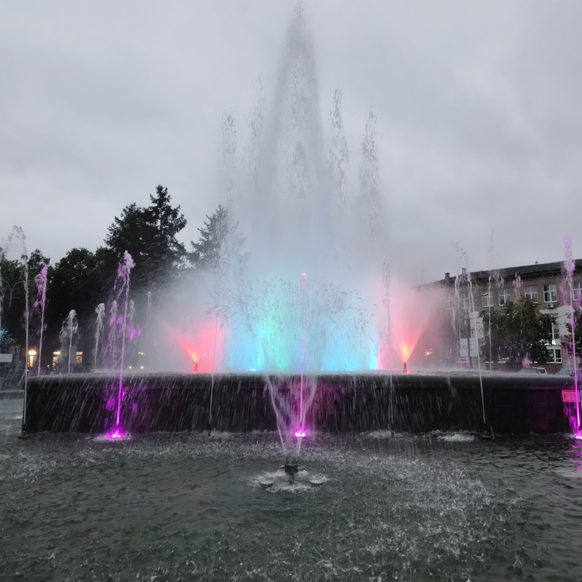 The image captures a large, illuminated fountain in the city of Chaikovsky, Russia. The fountain is the central subject, with water shooting upwards and outwards in various patterns. The water jets are lit with vibrant pink, blue, and green lights, creating a colorful spectacle against the overcast sky. In the background, there are dark green evergreen trees and a brick building with multiple windows. The water in the fountain's basin is turbulent, with many small splashes and ripples, indicating the force of the water jets. The lighting suggests it is either dusk or a very cloudy day, as the artificial lights of the fountain are prominent.