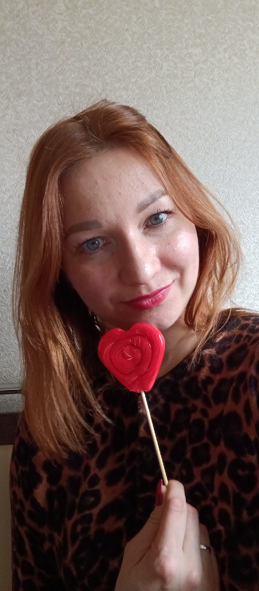 A woman with fair skin, light reddish-blonde hair, and bright blue eyes is depicted in a medium close-up, looking directly at the camera with a gentle smile. She is wearing a dark brown and black leopard print top. Her lips are adorned with red lipstick, matching the glossy red heart-shaped lollipop she holds in her right hand. The lollipop, which has a swirling pattern on its surface, is positioned below her chin, near her shoulder. Her right hand, with dark red painted fingernails, grasps the wooden stick of the candy. A silver-colored ring is visible on her left ring finger. The background is a plain, light beige-colored wall with a subtle textured pattern. There is no visible text in the image.
Location context: Unknown, Unknown