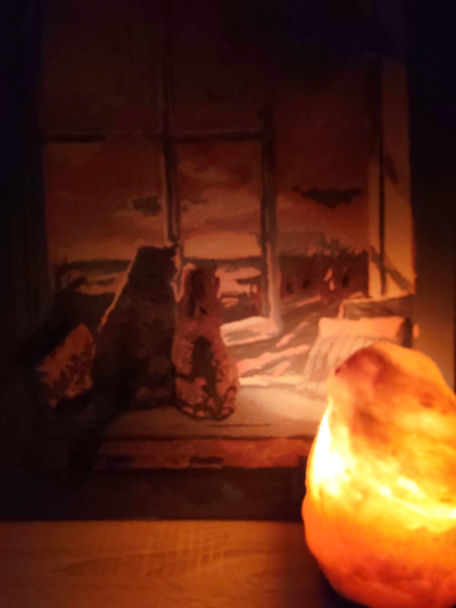 A dimly lit image depicts a painting next to a salt lamp. The painting features what appears to be two cats sitting on a windowsill, looking out at a landscape. The background is blurred but shows hints of fields and a cloudy sky. The salt lamp, located to the right of the painting, provides a warm, orange glow illuminating the lower right portion of the scene. The image is taken indoors, possibly in a room with dark walls. There are no people visible in the photo. The location in the city of Chaikovsky, Russia is not discernible from the image.