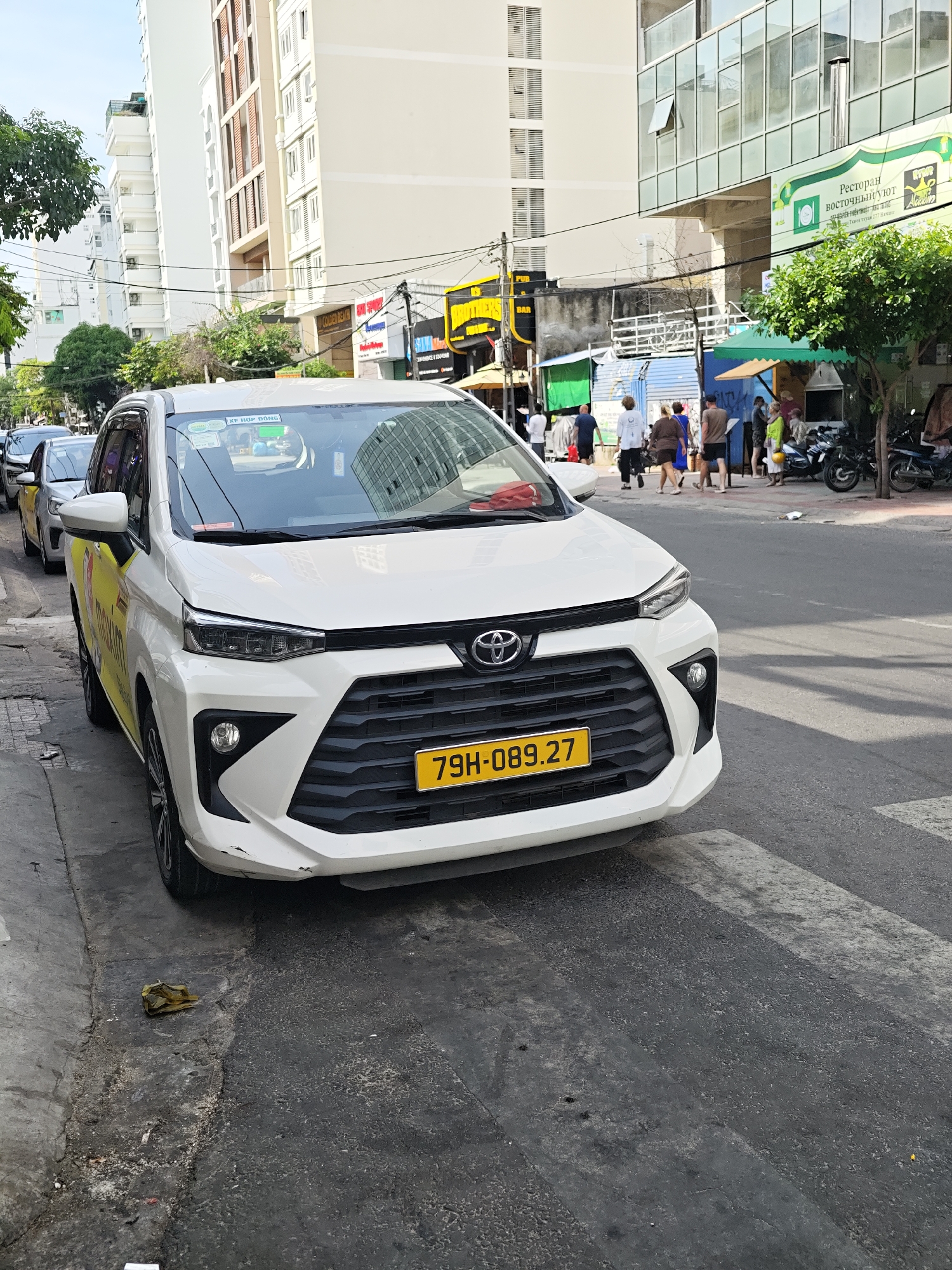A white Toyota Avanza taxi is parked on the side of a city street in Nha Trang Ward, Vietnam. The taxi has a yellow stripe along its side and a license plate reading "79H-089.27". In the background, there are tall buildings, other parked vehicles, trees, and several pedestrians walking on the sidewalk and near street vendors. The street is paved with asphalt and a pedestrian crossing is visible in the foreground.
