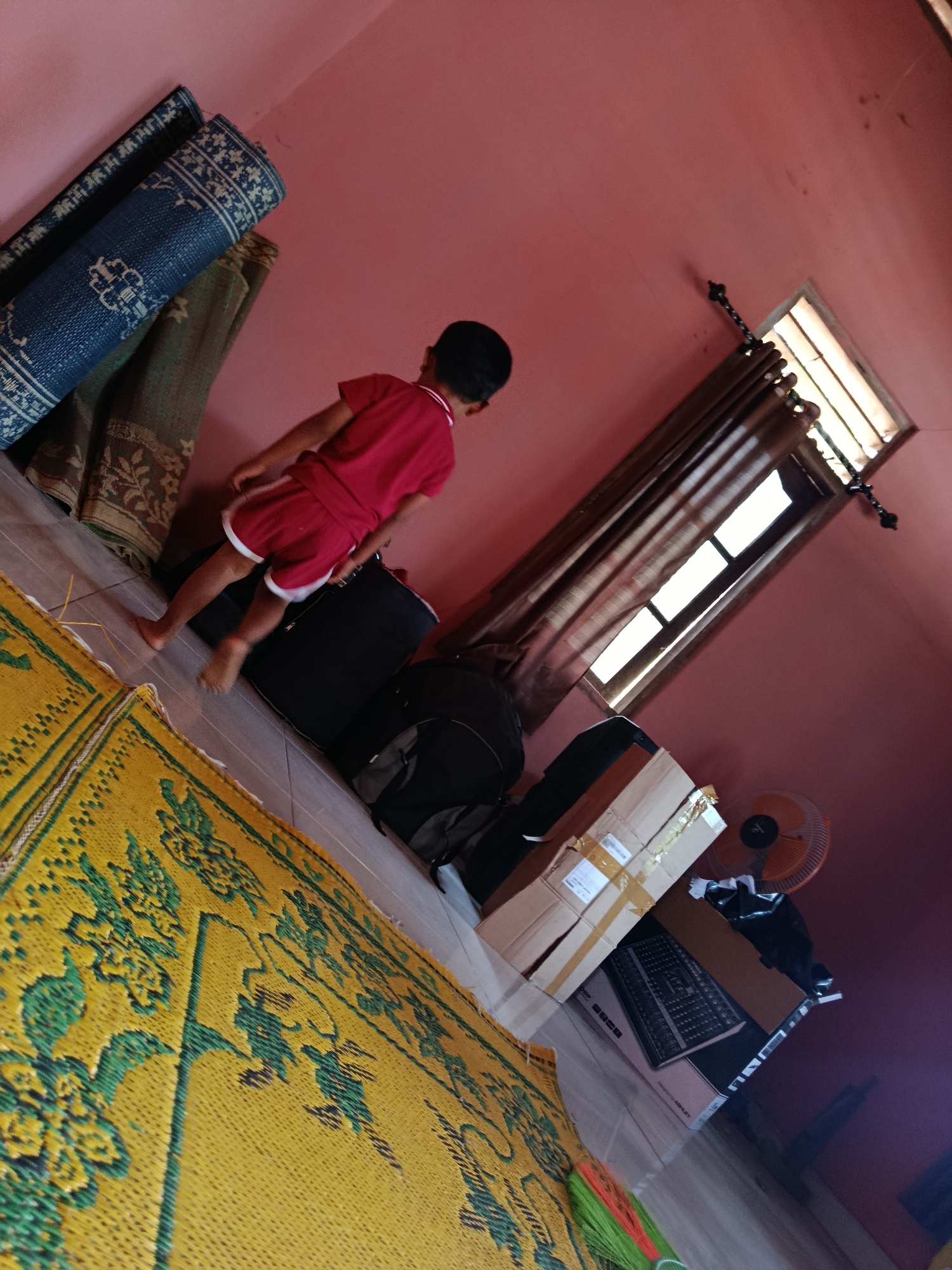 In a room with pink walls in Sedayu, Indonesia, a young child wearing a red t-shirt and shorts walks across a yellow mat with green floral patterns. Several rolled-up rugs are propped against the wall to the child's left, one dark blue with white intricate patterns, and another with earthy tones and floral designs. In the background, a window with brown curtains is visible, and a small orange electric fan stands near stacked cardboard boxes. The floor appears to be tiled.