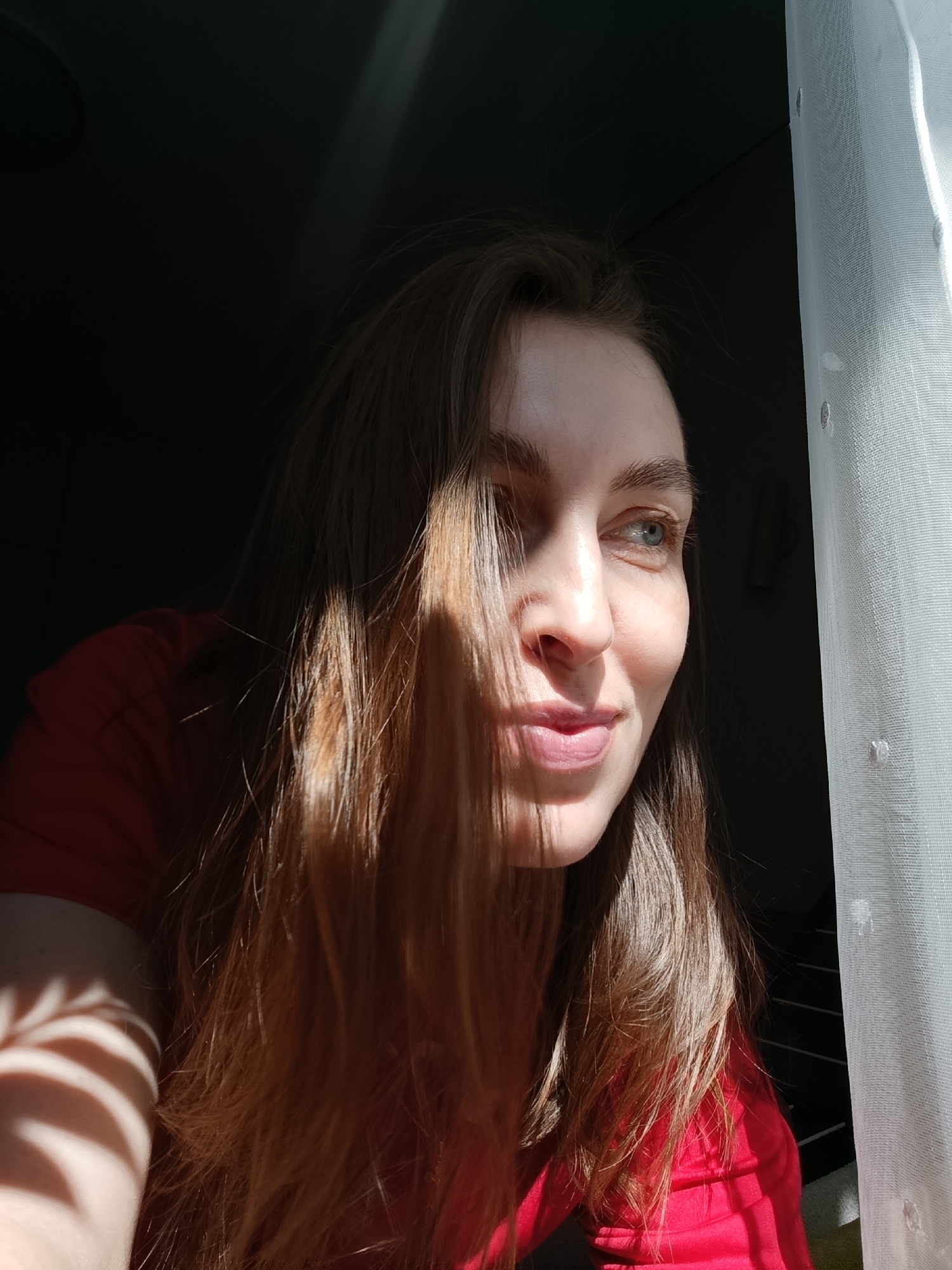 A young woman with long, light brown hair and blue-green eyes is depicted indoors, illuminated by a strong, directional stream of natural sunlight. She wears a red top and is positioned near a window, with a sheer white curtain visible along the right edge of the frame. Her head is turned slightly to the right, and her gaze is directed outwards or upwards, suggesting observation or contemplation, accompanied by a gentle smile.
The lighting creates a high-contrast effect, with intense sunlight brightly illuminating the right side of her face, her hair, and her shoulder, while the rest of the room and parts of her figure are cast in deep shadow. Distinct shadow patterns, appearing as stripes or slats, are visible on her left arm and forehead, indicating the sunlight is filtering through a patterned obstruction before reaching her. The background of the room is largely dark, enhancing the focus on the woman and the light. The scene conveys a sense of peacefulness or quiet enjoyment of a sunny moment. The strong, direct light suggests the time of day is either morning or late afternoon. The setting is a domestic interior, possibly in Chaikovsky, Russia. No visible text is present in the image.