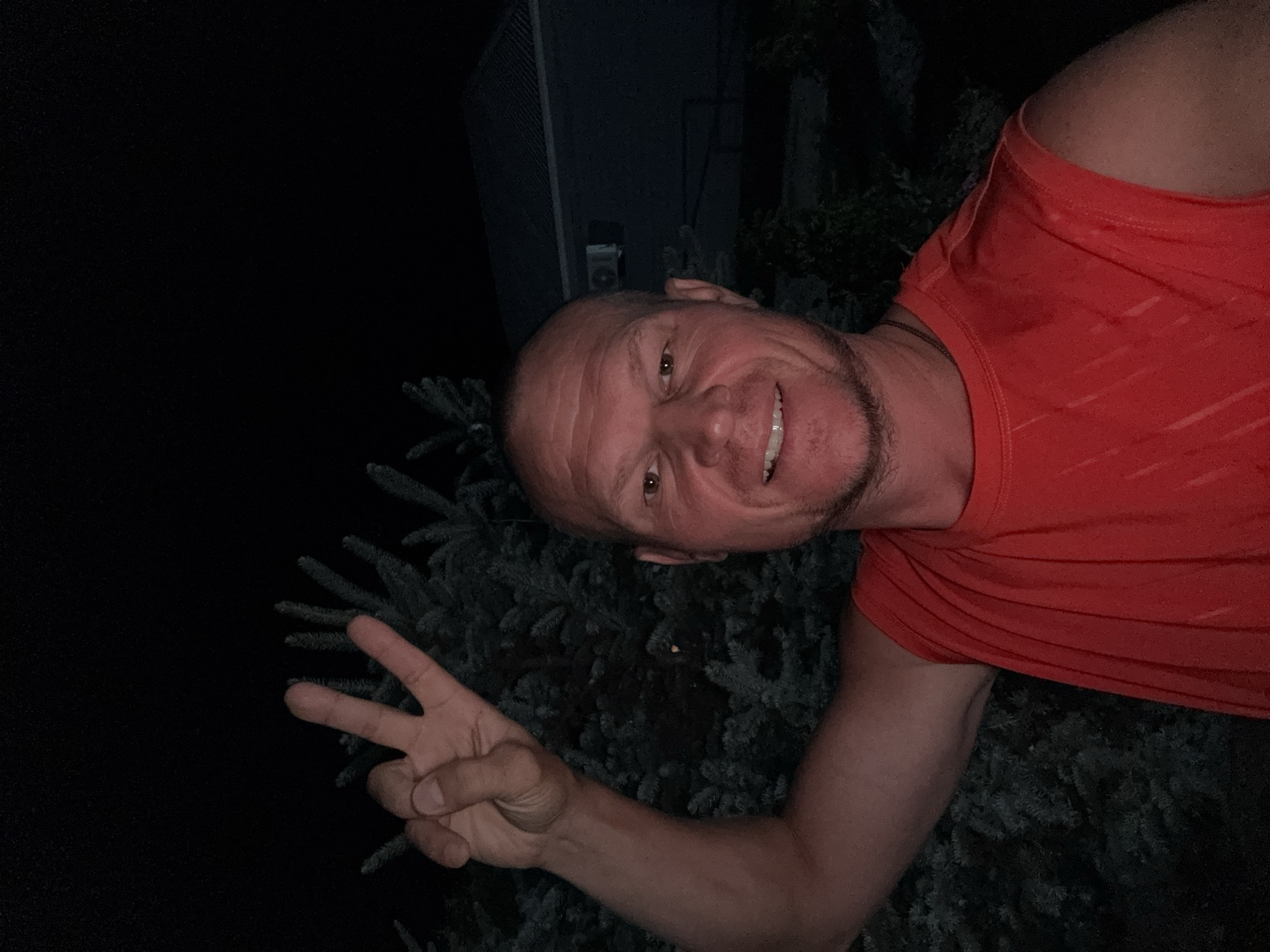 A bald, fair-skinned man with a light beard and a red sleeveless t-shirt is taking a selfie outdoors at night. He is smiling and holding up two fingers in a peace sign with his left hand. Behind him, dark evergreen trees are visible, and further in the background, a dark house with a light-colored roof and an air conditioning unit on the wall can be seen. The scene appears to be in the city of Urdzhar, Kazakhstan.