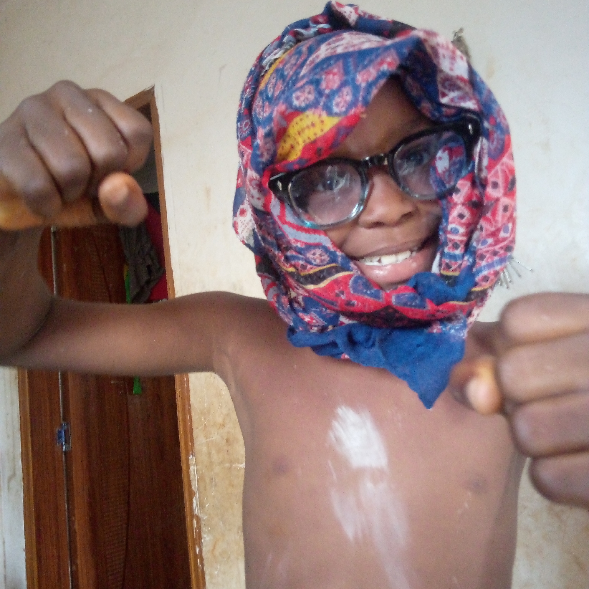 A dark-skinned child, appearing to be male, is centered in the frame. The child is shirtless and has a colorful patterned headscarf and black framed glasses. White powder is visible on the child's chest. The child's fists are clenched and held up towards the camera. Behind the child is a partially visible brown door. The location is Ogidi, Nigeria.