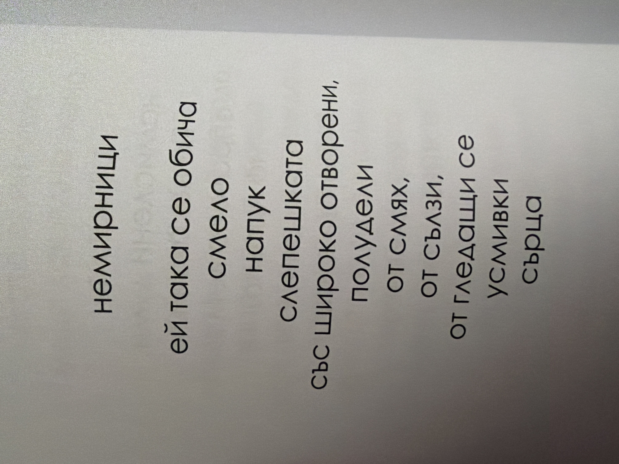 The image displays a close-up view of text printed on a light-colored paper, likely a page from a book or document. The text is in Bulgarian and is centered on the page. The font is sans-serif and appears to be in a bold or heavy weight. The lighting suggests an indoor environment with a slight shadow or gradient visible on the right side of the image, indicating the paper is not perfectly flat or is lit from the side. There are no people, objects, or environmental features other than the text and the paper itself visible in the frame. The text is arranged in a poem or verse format, with varying line lengths. The content of the text translates to: "restless ones, she loves so bravely, a brave, a departure, blind with wide open, half-crazy, from laughter, from tears, from watching, smiles, hearts." There is no information within the image that would allow for the identification of a specific city, location, or country like Pazardzhik, Bulgaria.