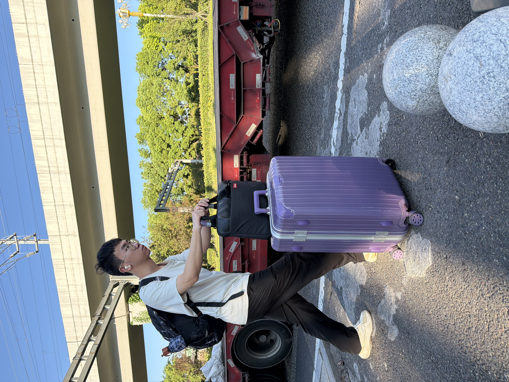 A young East Asian man wearing glasses, a white t-shirt, and black pants walks across an asphalt road. He carries a black messenger bag in his right hand and a large, purple suitcase with four wheels is positioned beside him. He has a backpack on his shoulders with an umbrella sticking out of a side pocket. Behind him, a red train car is visible, along with the underside of an elevated highway. The scene appears to be set in an urban area of Jiangjin, China, with trees and a blue sky in the background. Two large, round grey stones are in the foreground.