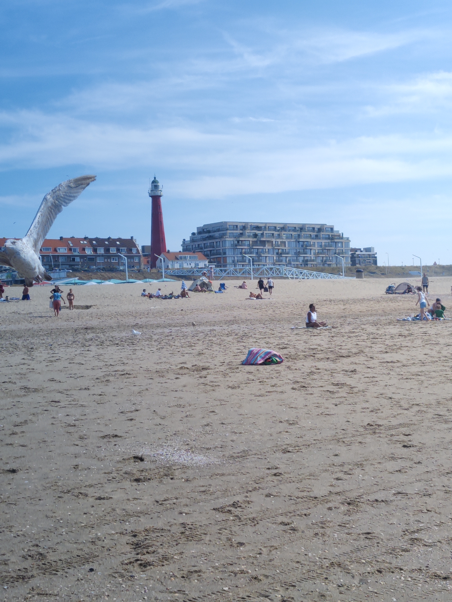 A wide shot captures a bustling beach scene with a prominent red and white lighthouse in the background. The foreground is dominated by sand, with scattered shells and tire tracks. Several beachgoers are visible: some are seated on the sand, others are standing, and one person is in a meditative pose. A large seagull is in flight in the upper left quadrant of the image. The scene also includes a modern apartment building, other smaller buildings, beach tents, and umbrellas. The sky is a bright blue with scattered white clouds. This image was taken at Scheveningen beach in The Hague, Netherlands, a popular seaside resort town.