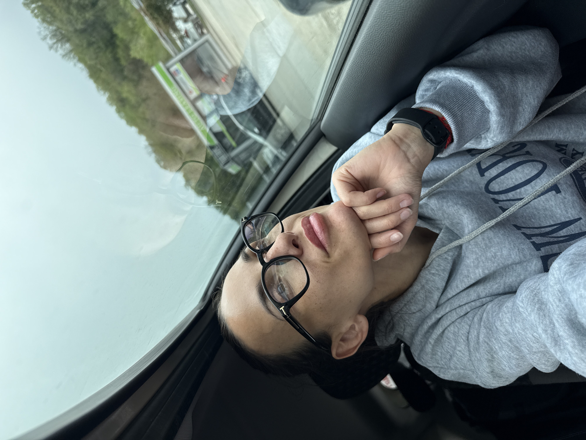 The image captures a diagonally oriented view of a person inside a vehicle, seen from a slightly elevated perspective. The subject is a young adult with dark hair, wearing black-framed eyeglasses that feature a 'T' logo on the temple arm. They are dressed in a grey hooded sweatshirt with visible drawstrings, which displays partially legible dark text, including "NO" and parts of other letters that resemble "YORK." A black digital wristwatch, revealing a red string bracelet underneath, is worn on their left wrist. The person's hands are clasped together, resting against their mouth and chin, as they gaze upwards with a contemplative or observant expression.
The setting is the interior of a car, indicated by the dark headliner, window frame, and pillar. Through the car's windows and reflections on the glass, an overcast sky and blurry outdoor elements are visible. These include green foliage and the reflection of a structure with some green-accented signage, suggesting an outdoor environment, possibly urban or suburban, during daytime. The overall impression is one of quiet repose or thought within a car on a cloudy day, located in Pleven, Bulgaria.
