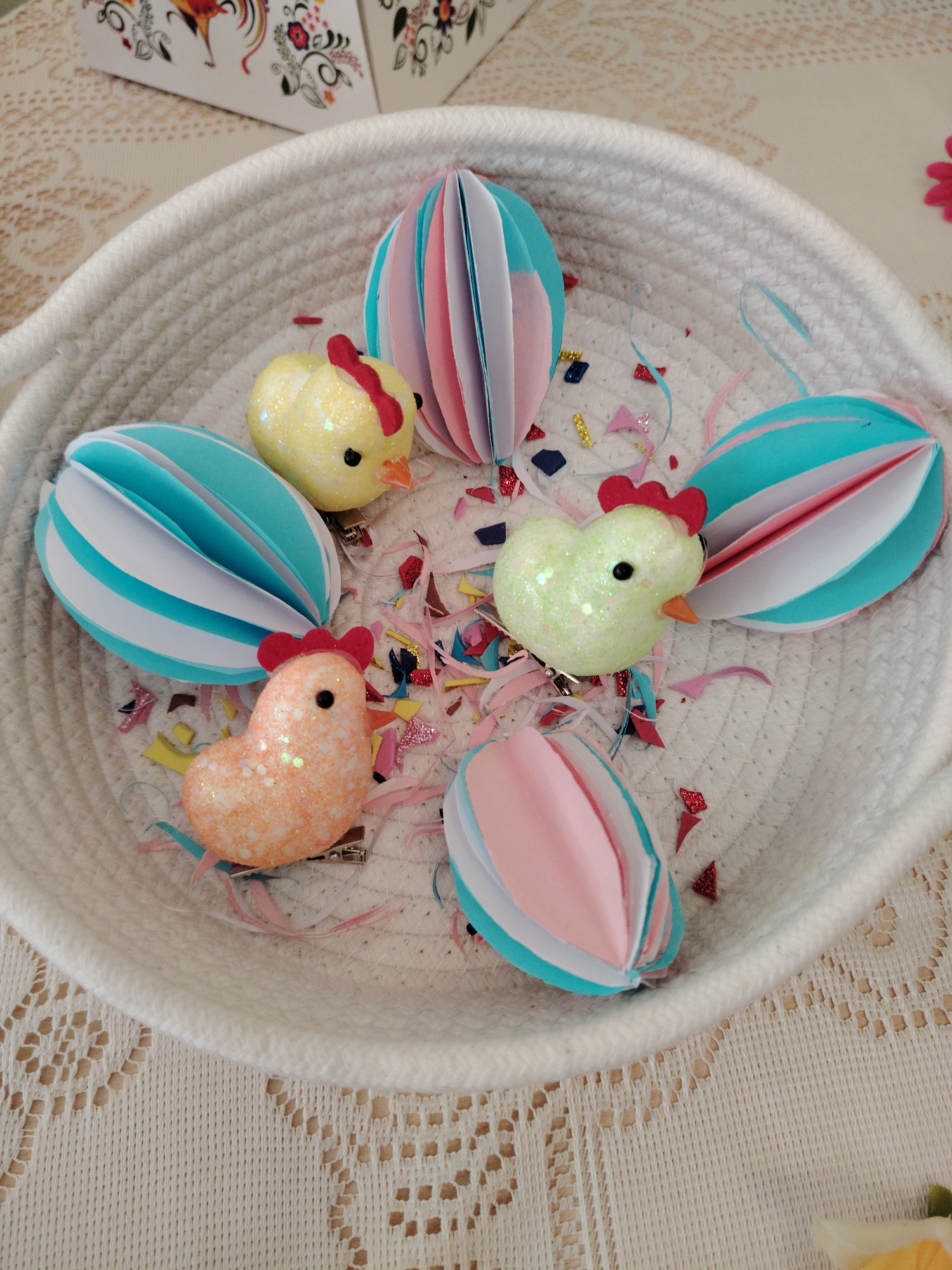 The image shows a white woven basket filled with Easter decorations. Inside the basket are three decorative paper eggs, each made from folded paper in blue, white, and pink. Interspersed among the eggs are three glittery, chick-shaped decorations in yellow, green, and orange. Small pieces of colorful confetti are scattered around the decorations. The basket sits on a tablecloth with a lace pattern. Part of a box with a rooster design is visible in the upper left corner of the image. The location in the city of Sarapul, Russia is unknown.