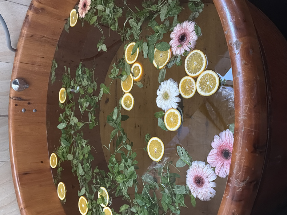 A large, circular wooden bathtub, constructed from vertical light-brown wooden staves, is filled with a dark amber liquid. Floating on the liquid are numerous green mint leaves, approximately 20-30 round citrus slices (predominantly orange), and five gerbera daisies (three pink, two white). A silver-colored metal faucet is mounted on the inner wall of the tub towards the upper center, and a grey corrugated pipe is visible entering the tub from the upper right side. The background indicates an indoor setting with a light-colored tiled wall. No individuals are present. This scene is located in Petah Tikva, Israel.