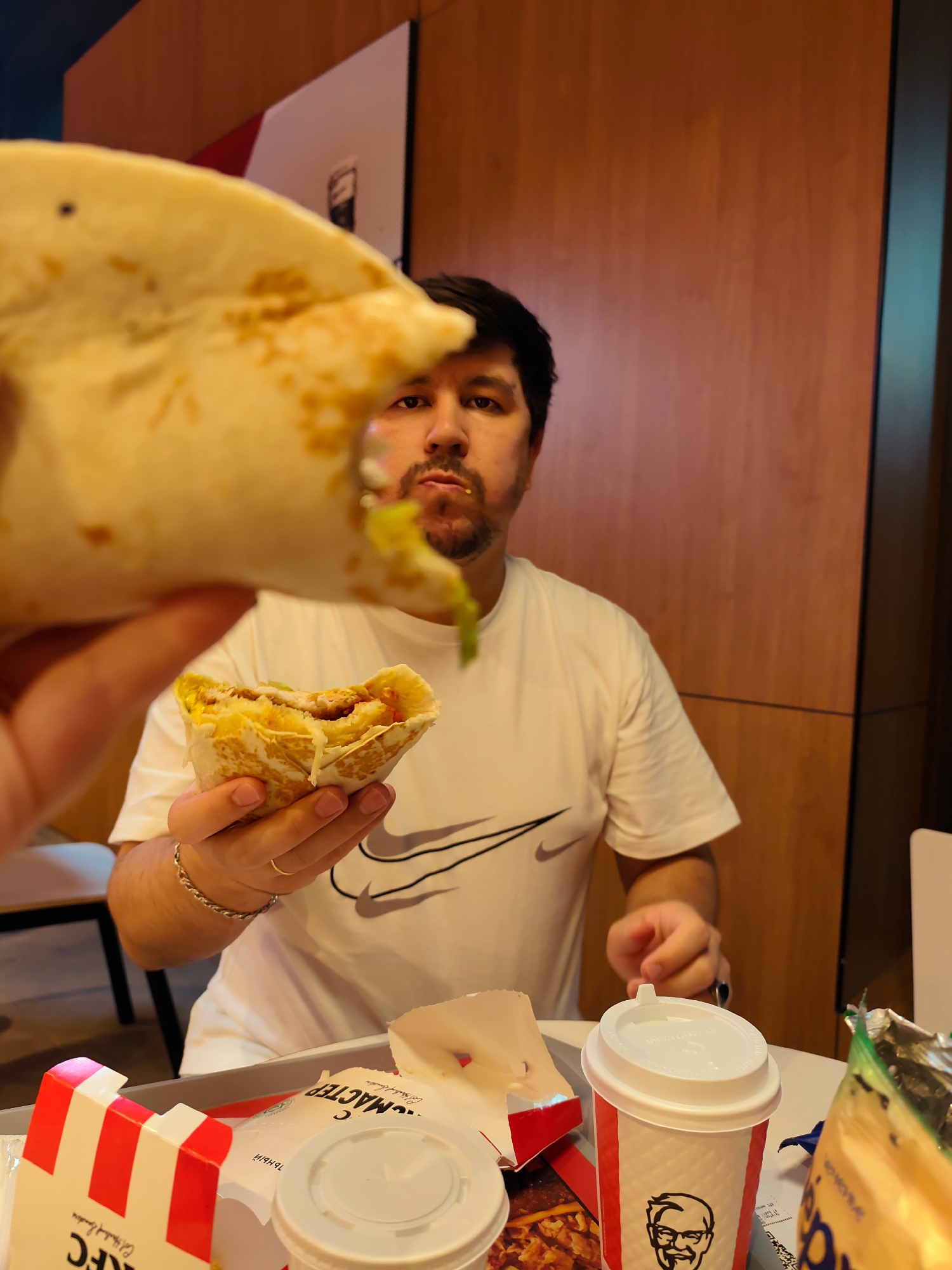 A man with dark hair and a beard is holding a halved wrap in his left hand and taking a bite out of another wrap in his right hand. He is wearing a white t-shirt with a grey Nike logo. In front of him is a KFC branded tray with a red and white striped box, a white lid cup, and a green and yellow bag. The background consists of wooden panels and a light-colored wall. The scene appears to be indoors, likely a fast-food restaurant in Fergana City, Uzbekistan.
