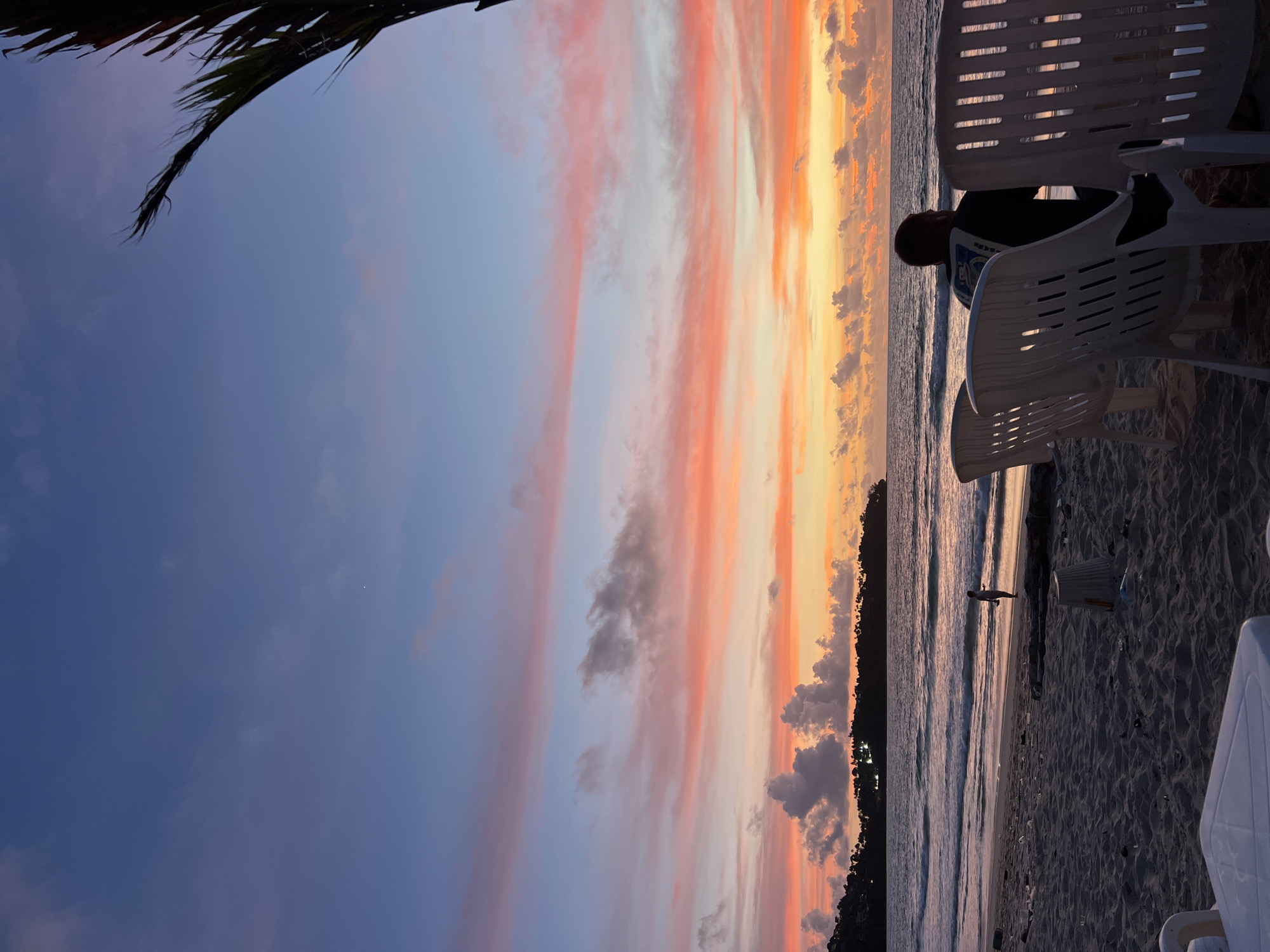 At sunset on a beach in Choeng Thale, Thailand, a lone individual stands near the water's edge, their silhouette small against the vastness of the ocean. In the foreground, several white plastic chairs are arranged on the sand, some occupied by a person facing the sea, while others are empty. The sand itself is littered with debris, including plastic bottles and other discarded items. A palm frond juts into the frame from the upper right corner, its dark silhouette contrasting with the vibrant hues of the sky. The sky is a canvas of color, with streaks of pink, orange, and blue clouds scattered across the horizon. In the distance, a tree-lined island with illuminated buildings marks the coastline. Waves gently lap at the shore, reflecting the warm light of the setting sun.